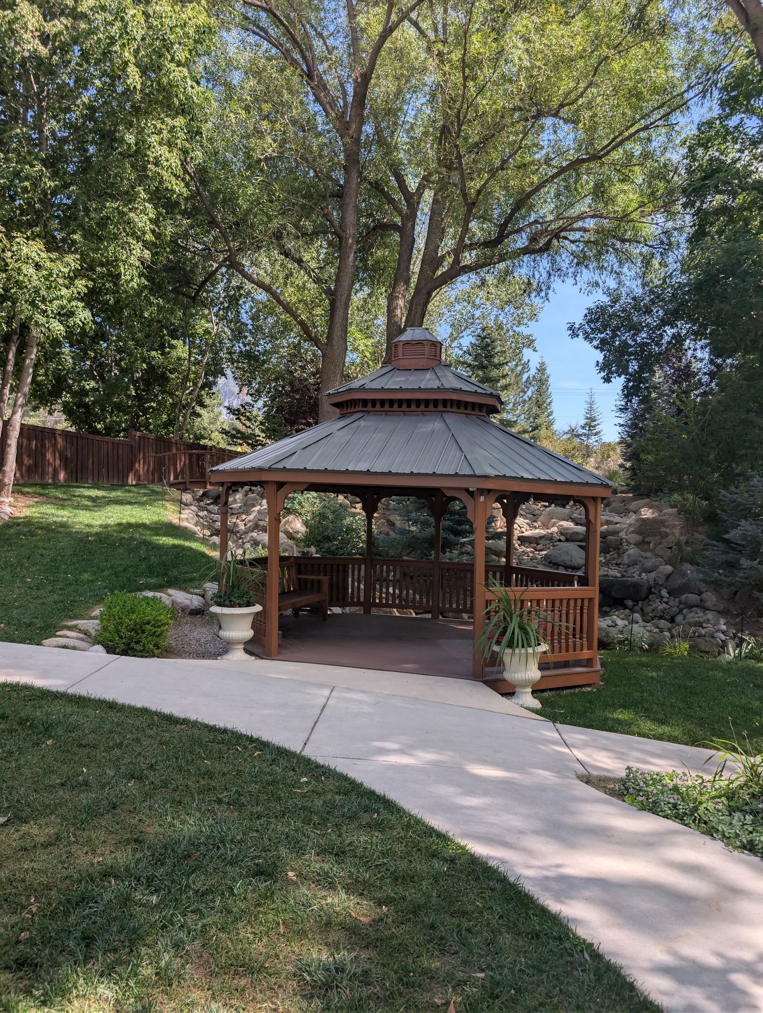 A brown wooden octagonal gazebo set against a rock-lined hillside waterfall feature, surrounded by mature trees