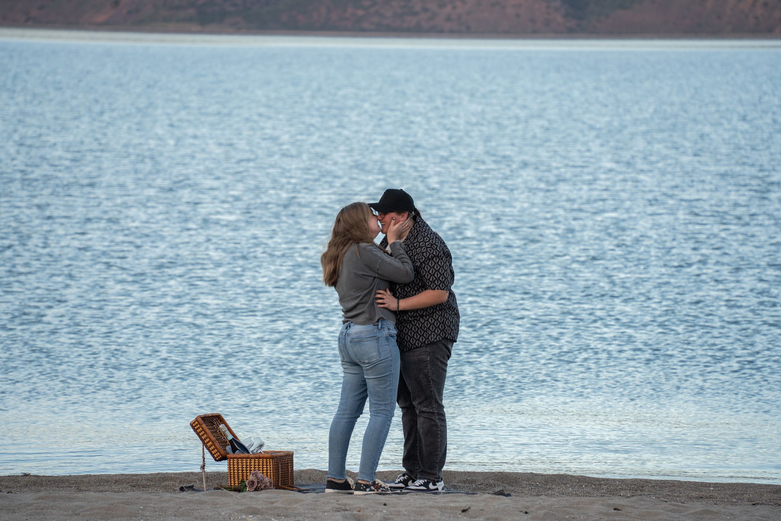 lesbian Couple sharing a romantic kiss on a sandy beach shore beside a picnic basket with calm blue water behind them.