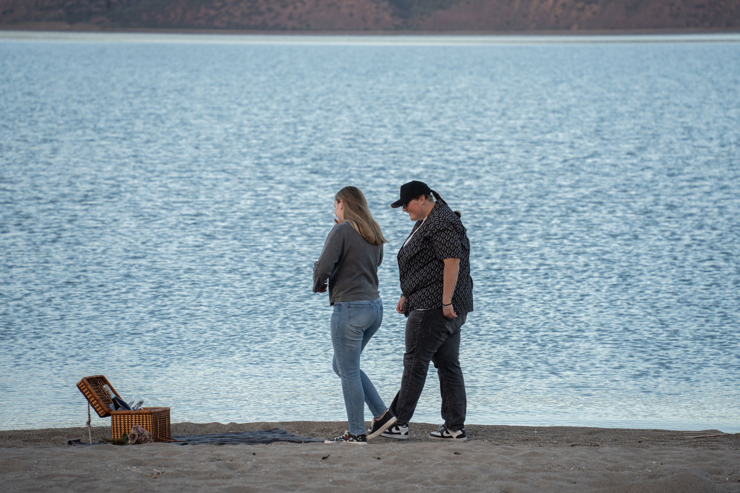 Couple walking along a sandy bear lake beach shore near calm blue water with a picnic basket nearby.