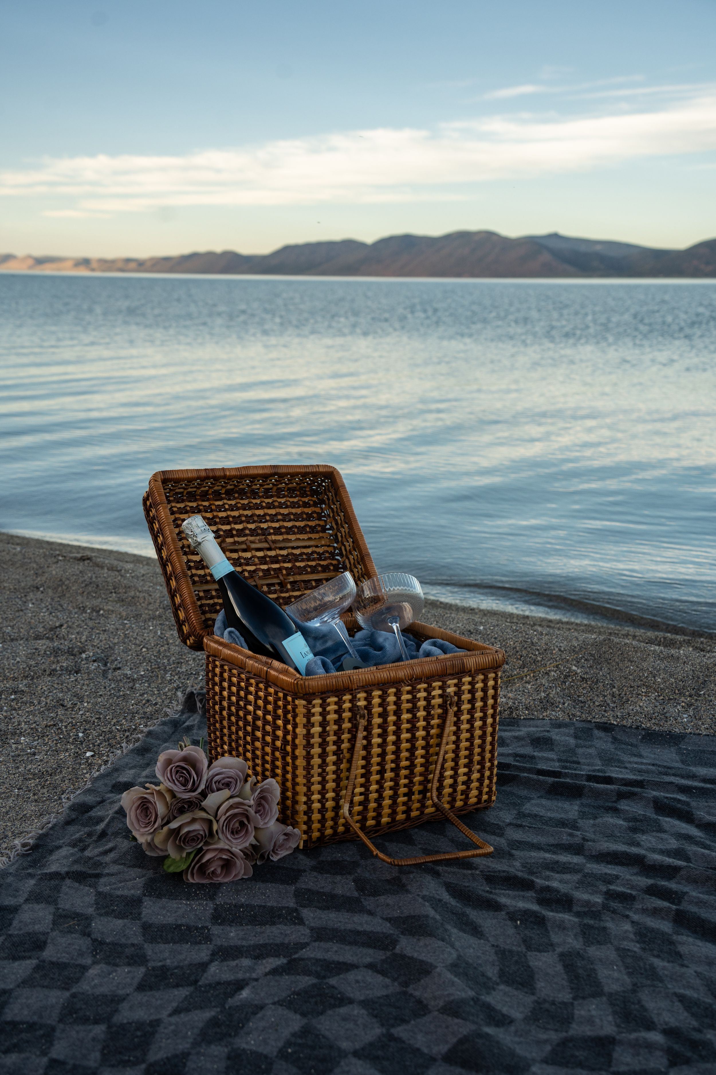 Romantic picnic basket with champagne and roses on a sandy beach at sunset with calm water and mountains in the background.