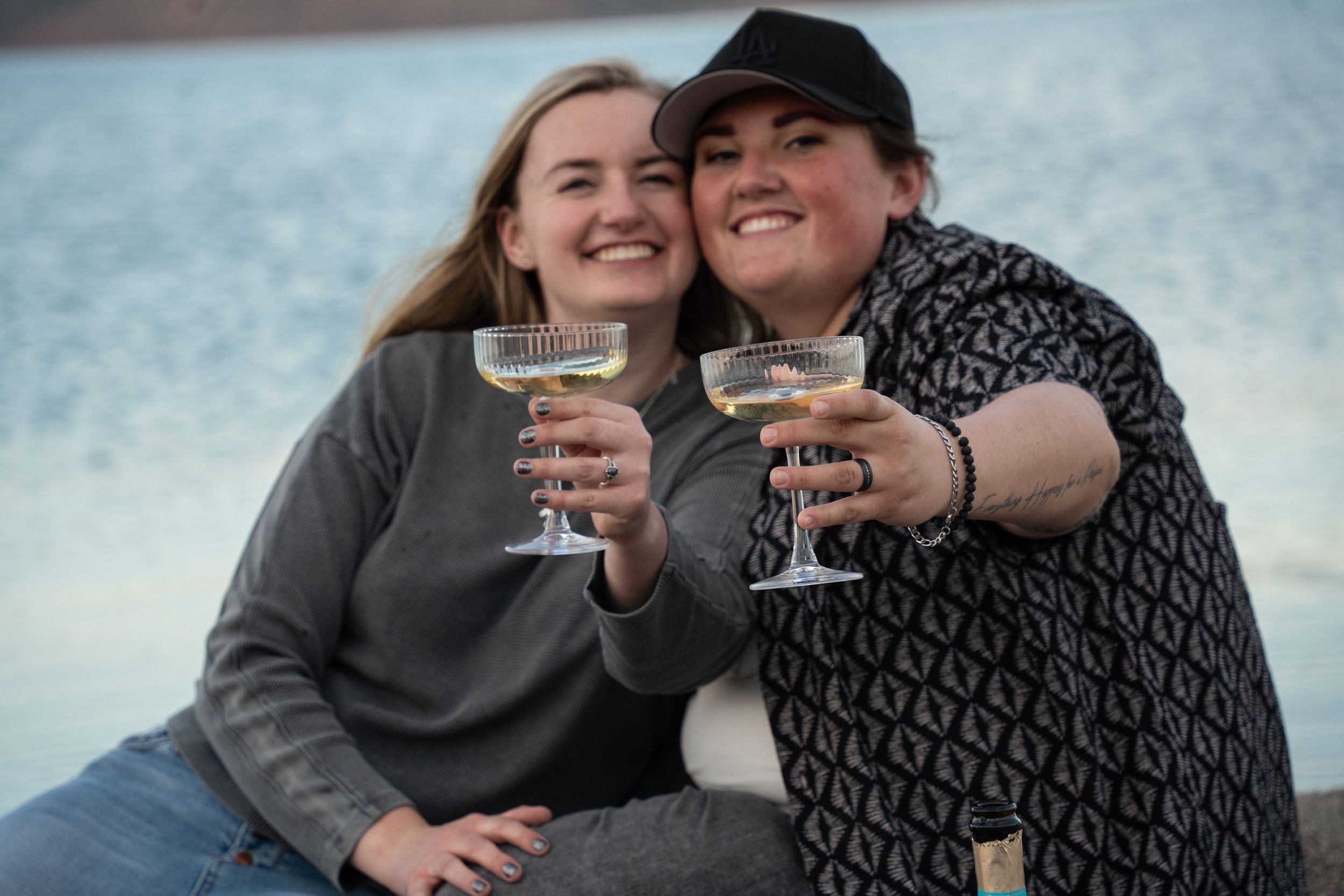wlw couple holding champagne glasses towards the camera and smiling together with blue water of bear lake behind them