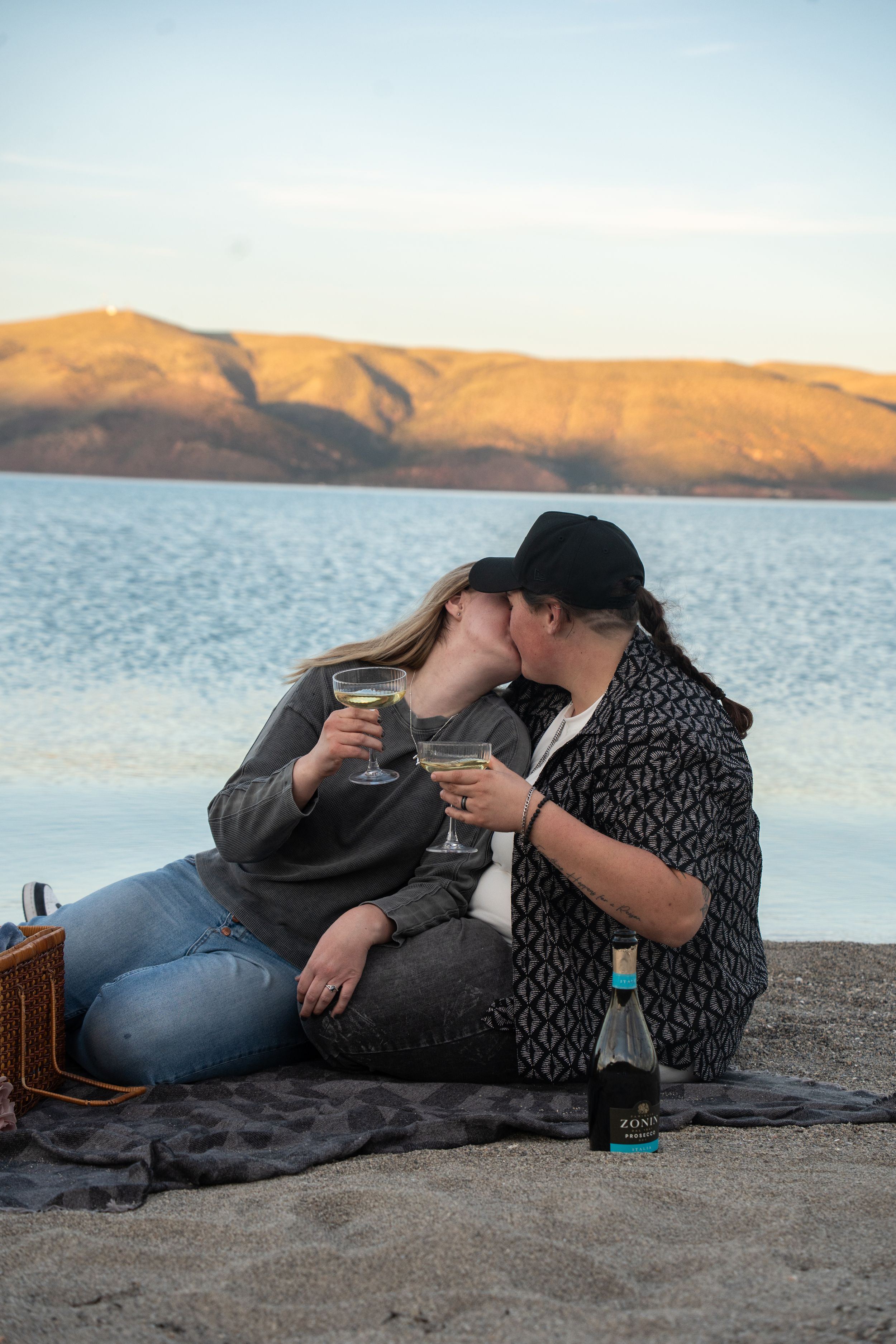 two women kissing on beach blanket at sunset, holding wine glasses with picnic basket and bottle nearby.