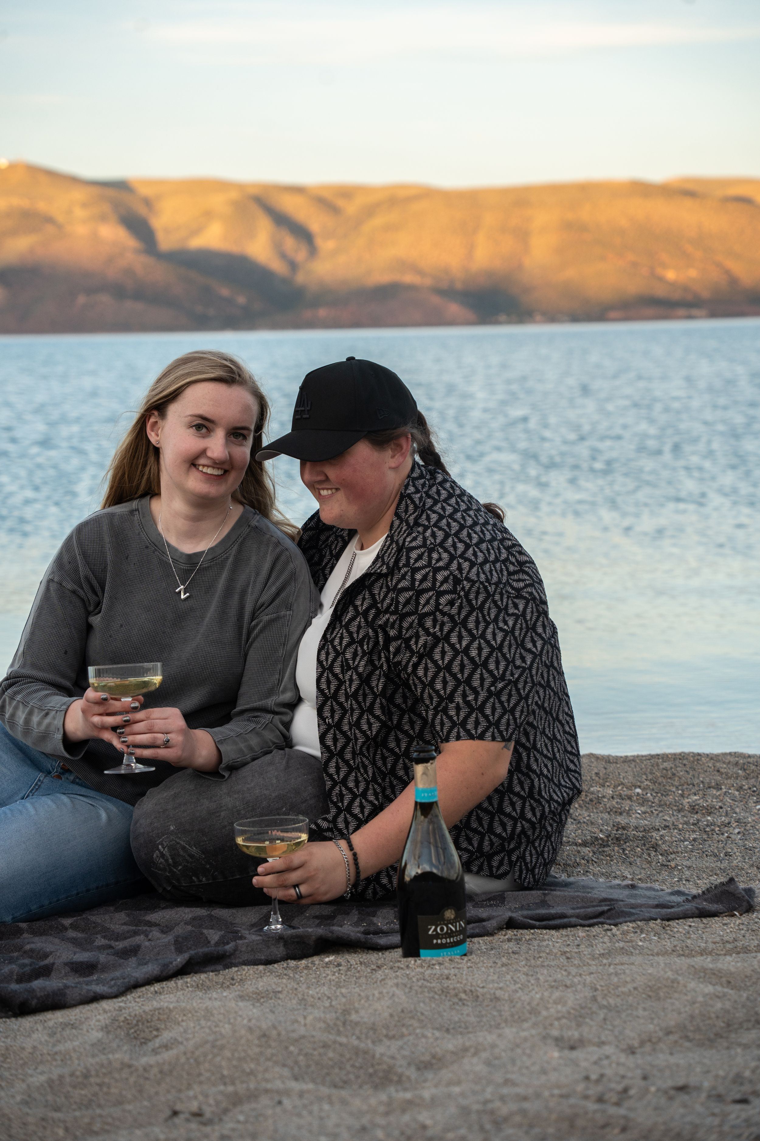 Two women sitting on a sandy beach sharing champagne, with a scenic lake and mountain backdrop at sunset.