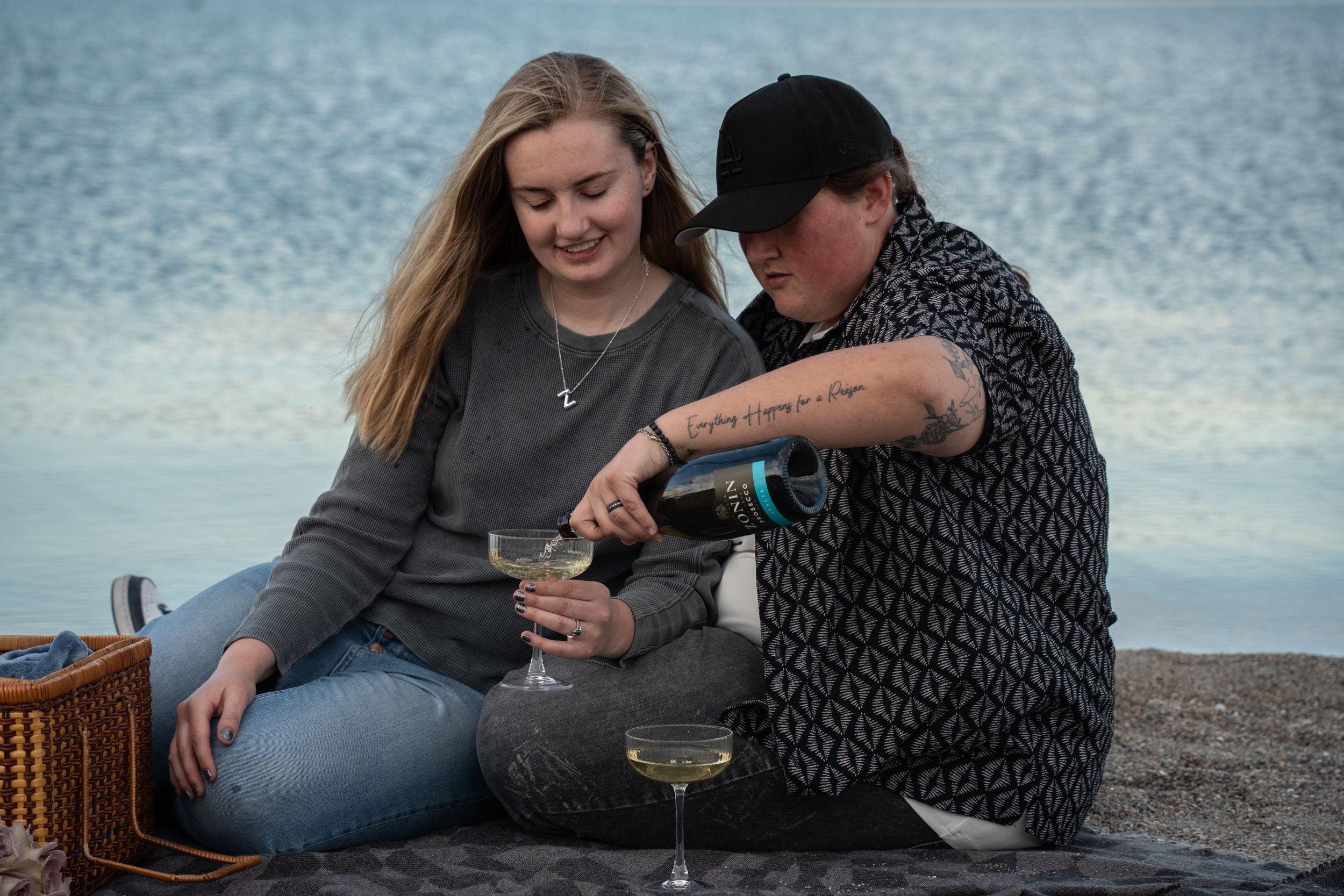 Couple pouring champagne on a beach blanket with a picnic basket, enjoying a romantic seaside date at dusk.