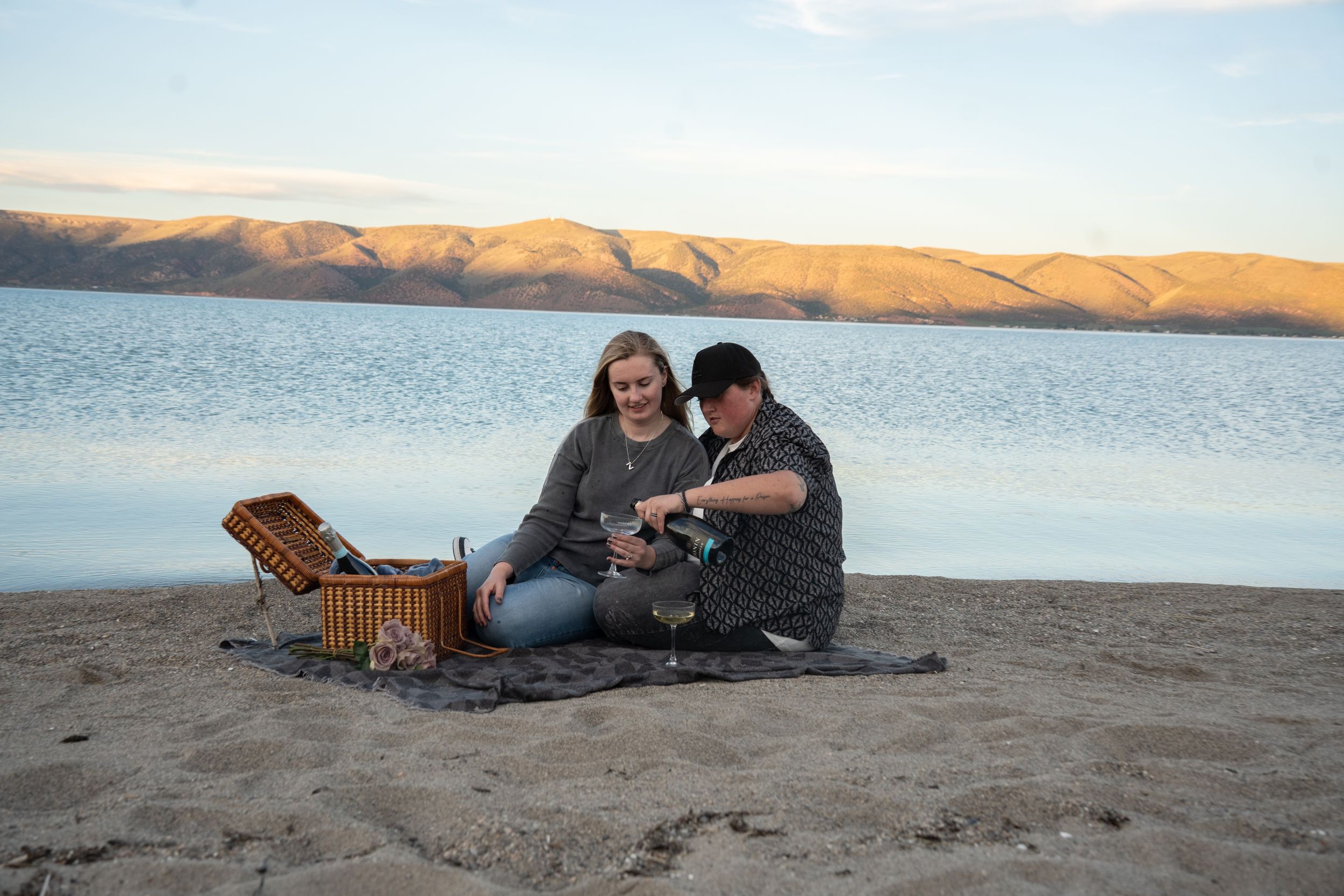engaged sitting on sandy beach blanket with picnic basket, enjoying lakeside view of bear lake with mountain backdrop at sunset.