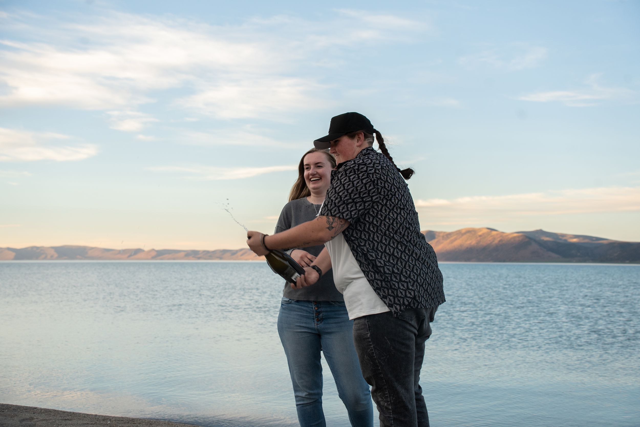 Couple laughing and holding a bottle of champagne on a lakeside beach with mountains and blue sky in the background.