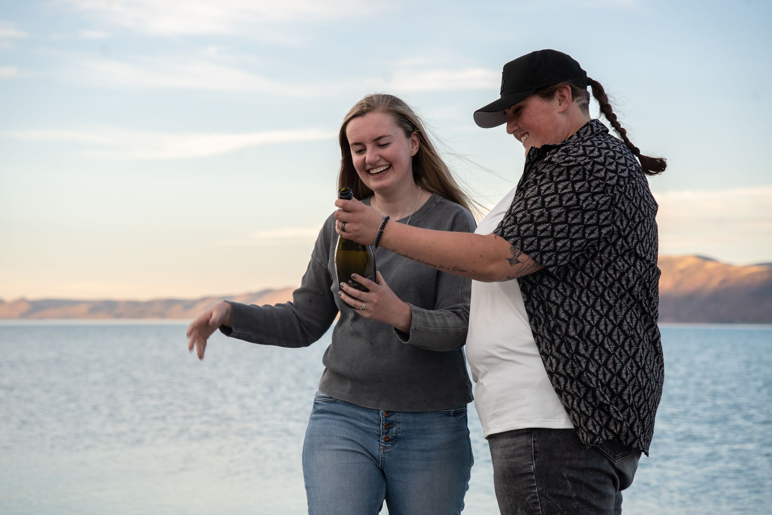 Two women laughing and sharing a bottle by a scenic lake with mountains in the background.