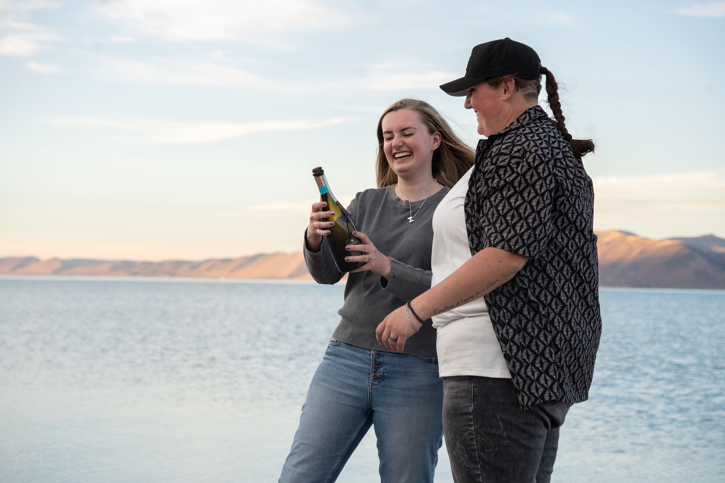 Two girlfriends laughing and looking at a bottle of champagne by a scenic lake with mountain views at sunset.