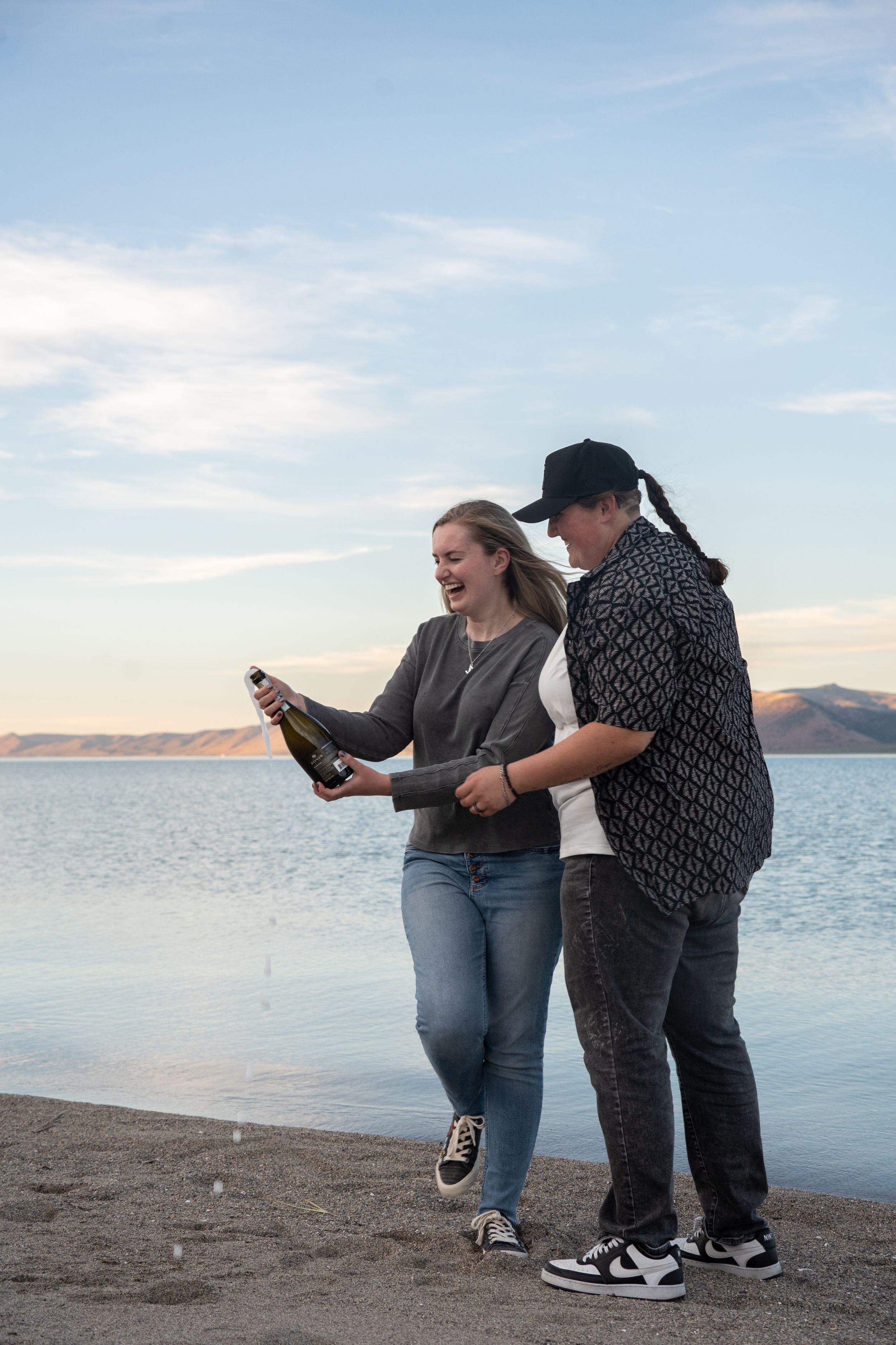 lesbian couple popping a bottle of champagne with champagne spilling everywhere and bear lake in the background