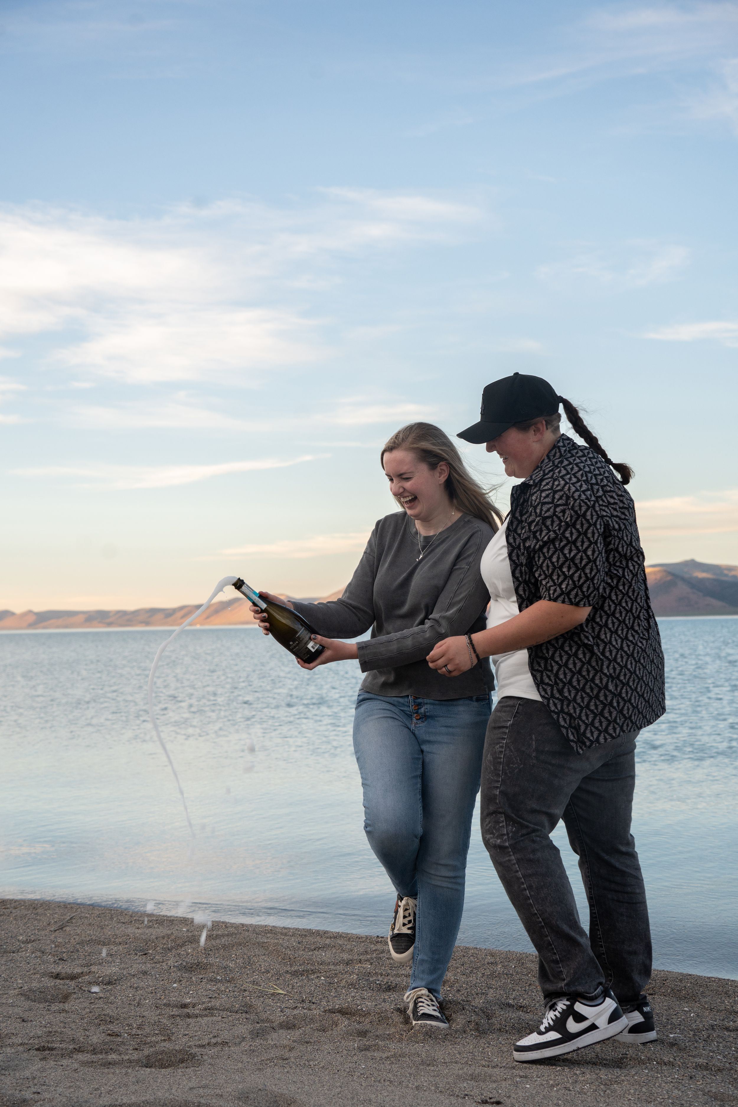 lesbian couple popping a bottle of champagne with champagne spilling everywhere and bear lake in the background