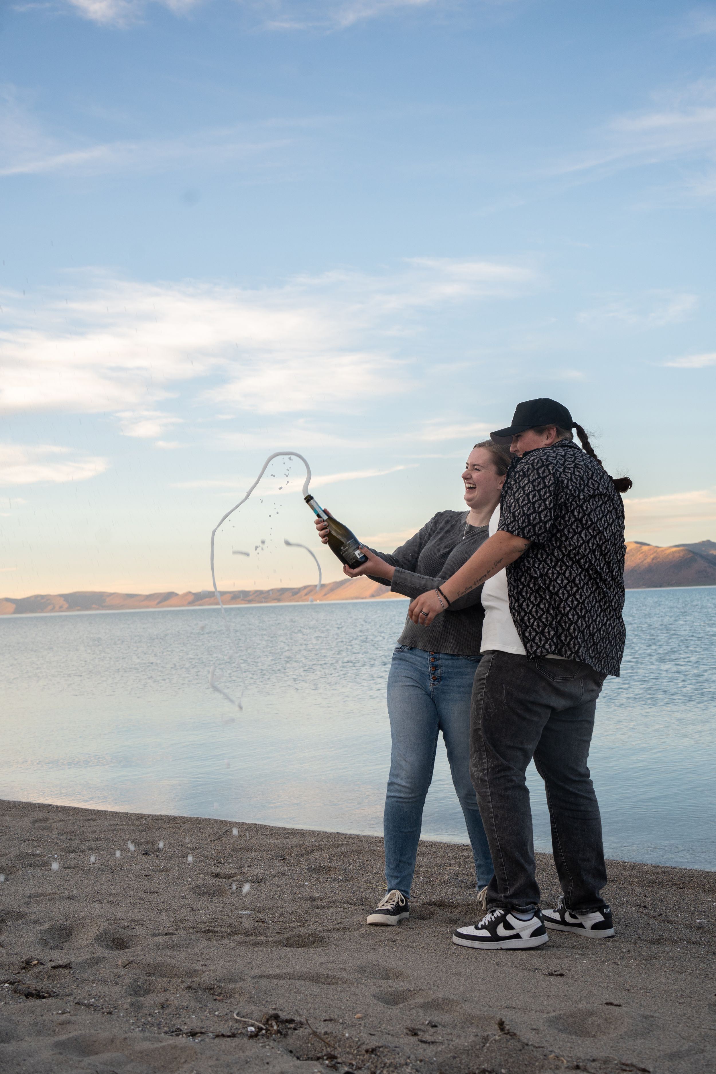 lesbian couple popping a bottle of champagne with champagne spilling everywhere and bear lake in the background