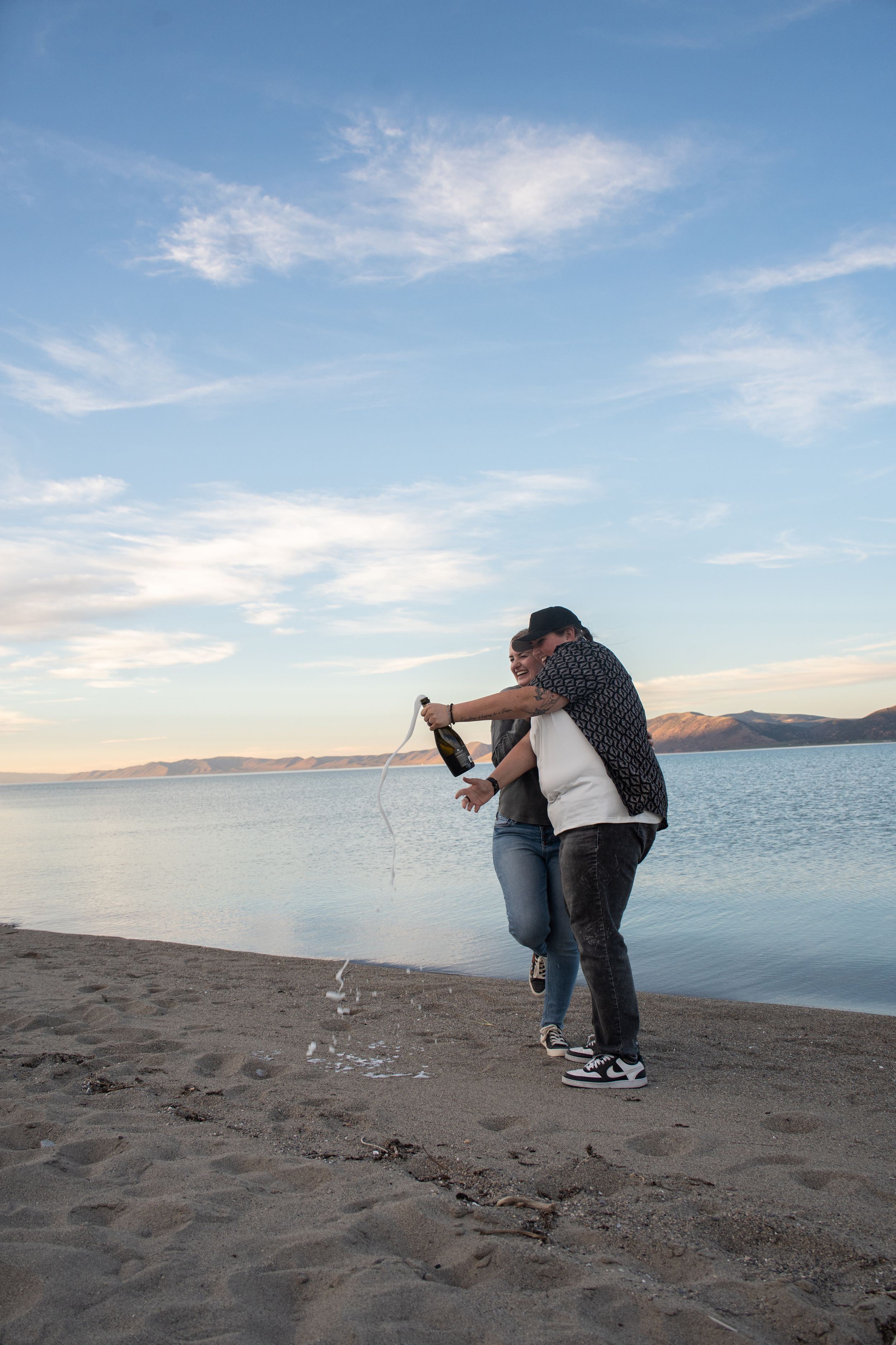 lesbian couple popping a bottle of champagne with champagne spilling everywhere and bear lake in the background