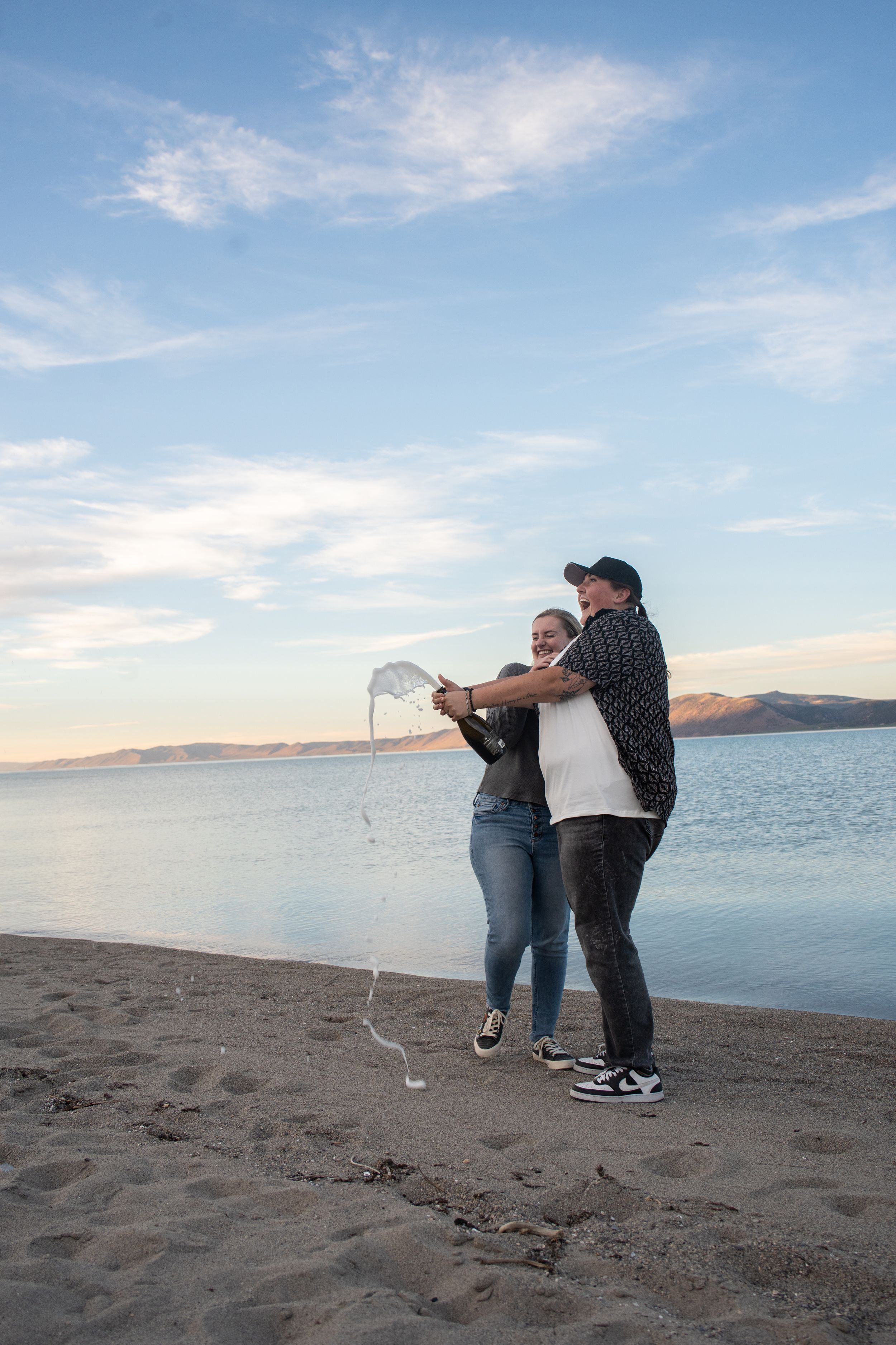 lesbian couple popping a bottle of champagne with champagne spilling everywhere and bear lake in the background