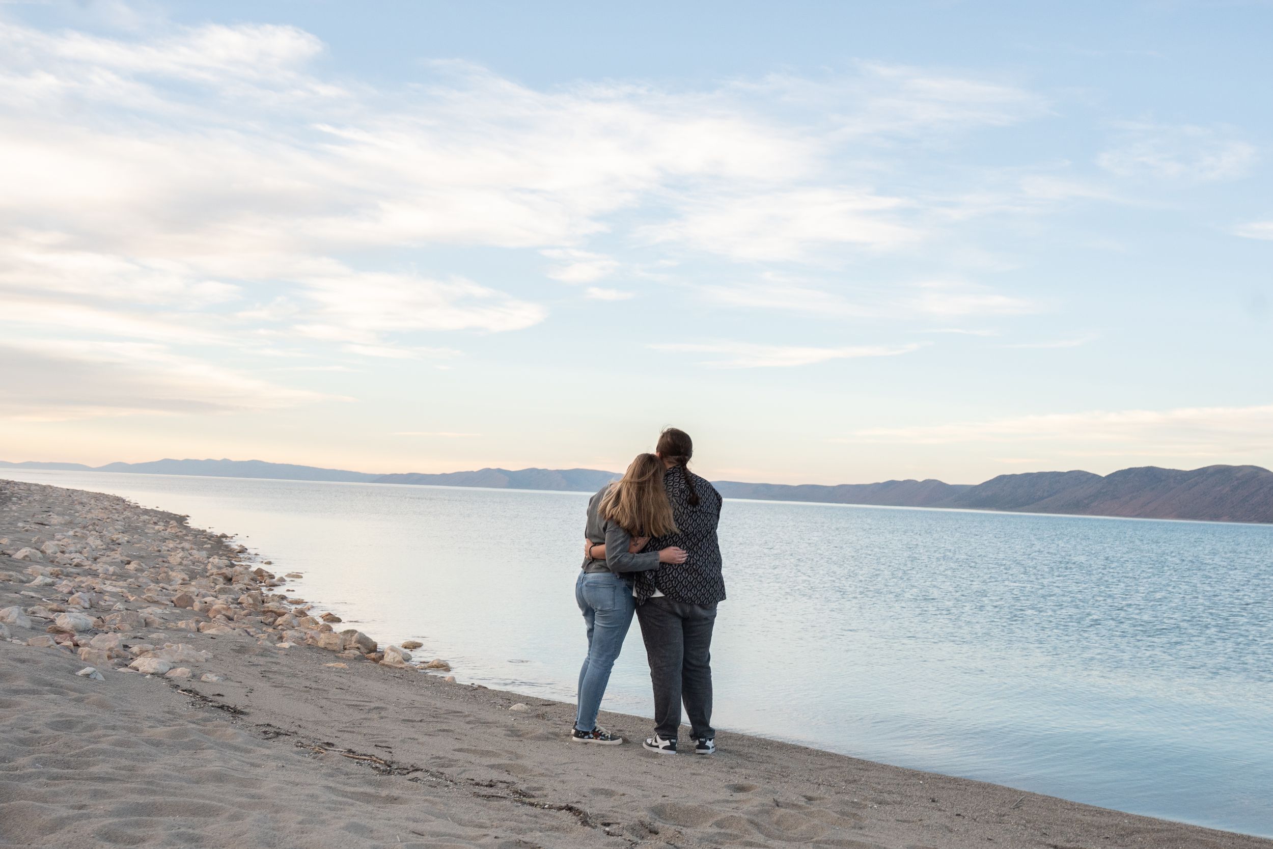 Two women embracing on a sandy beach shore at dusk with calm water and mountains in the background.