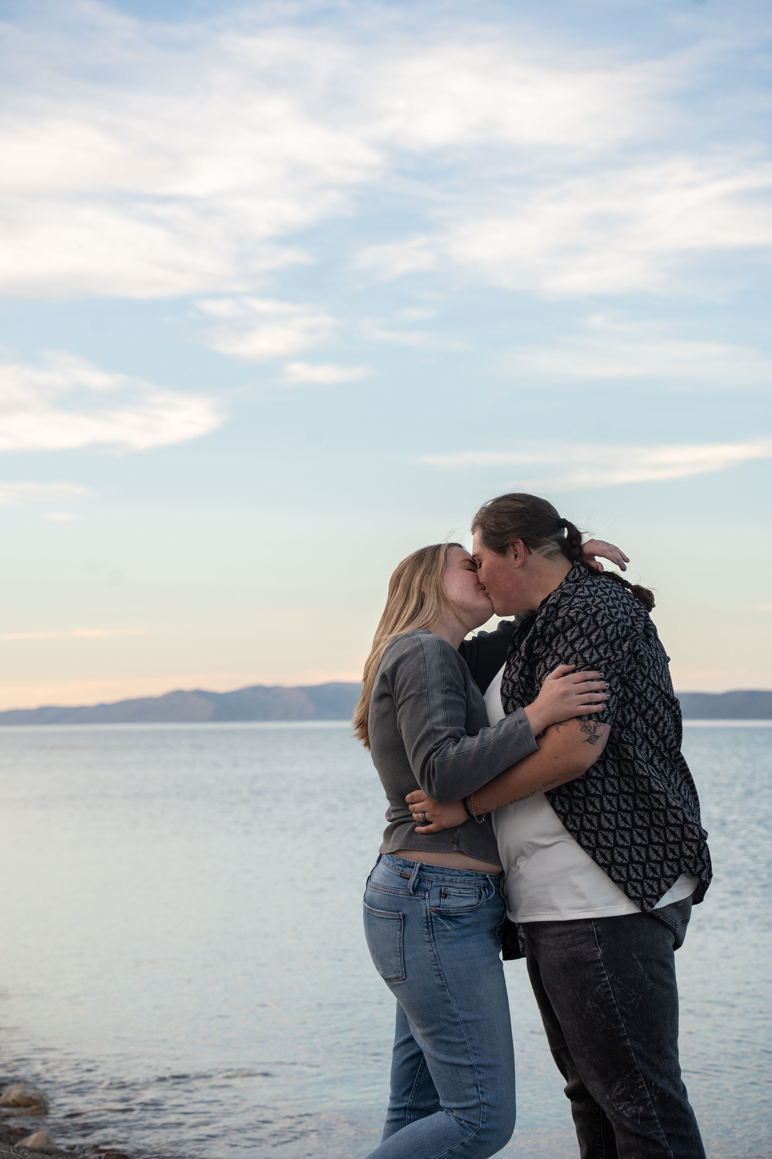 newly engaged Couple sharing a romantic kiss by the meal lake waterfront at sunset, with mountains and cloudy sky in the background.