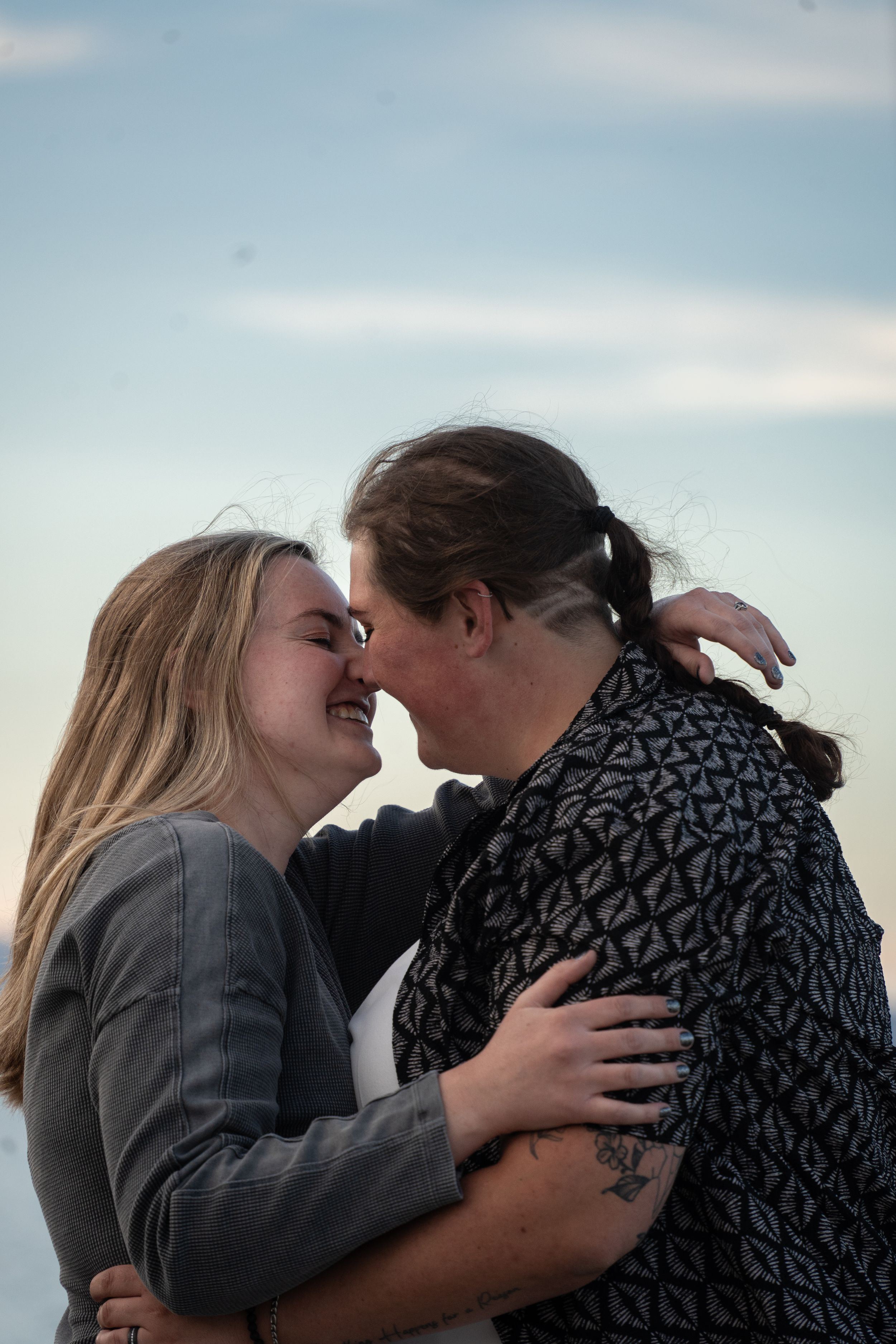 Two women embracing and sharing a tender moment outdoors against a blue sky backdrop.