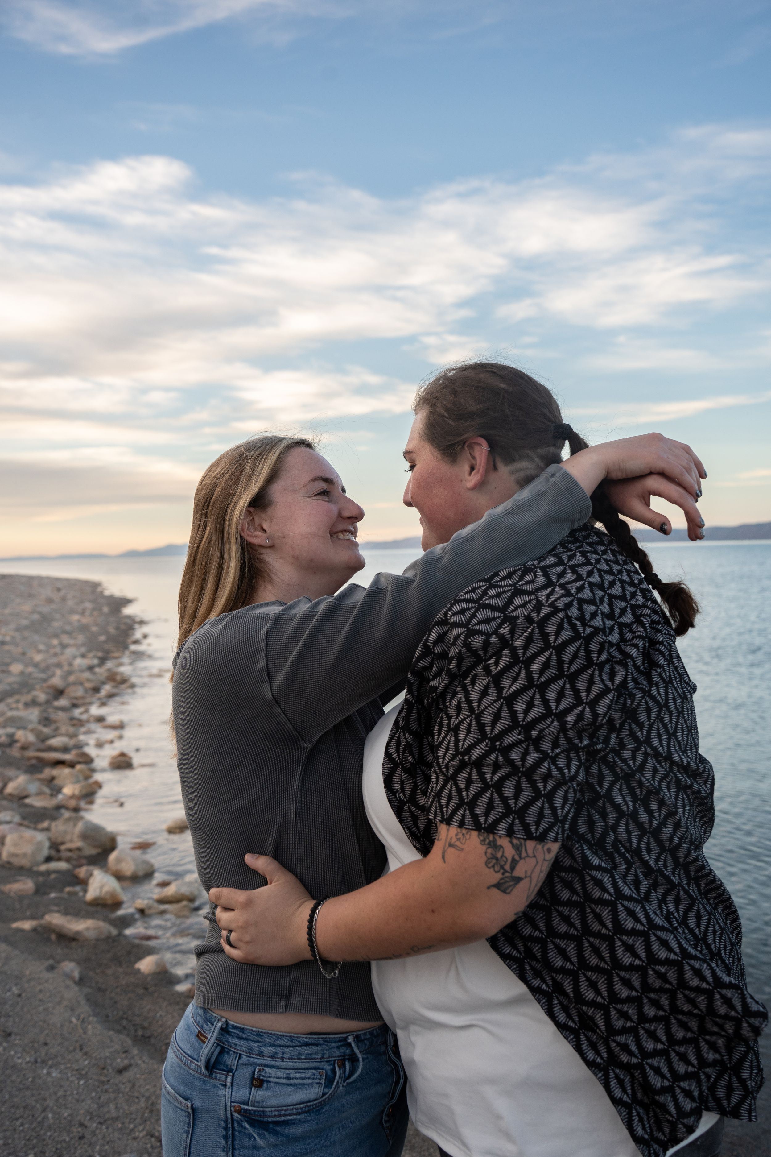 Two women embracing and smiling at each other on rocky bear lake beach at sunset.