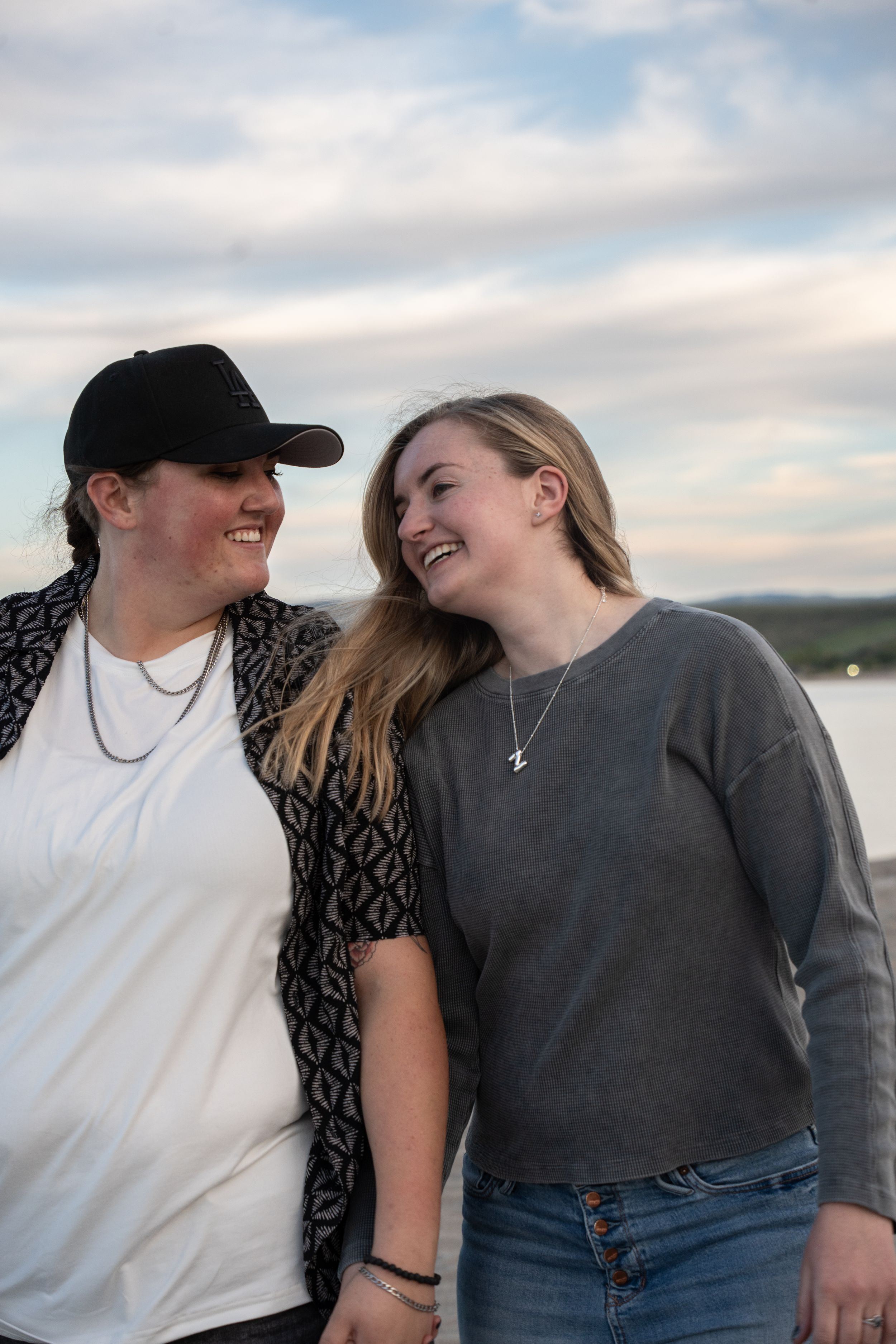 Two young women smiling and laughing together outdoors near water under a cloudy sky.