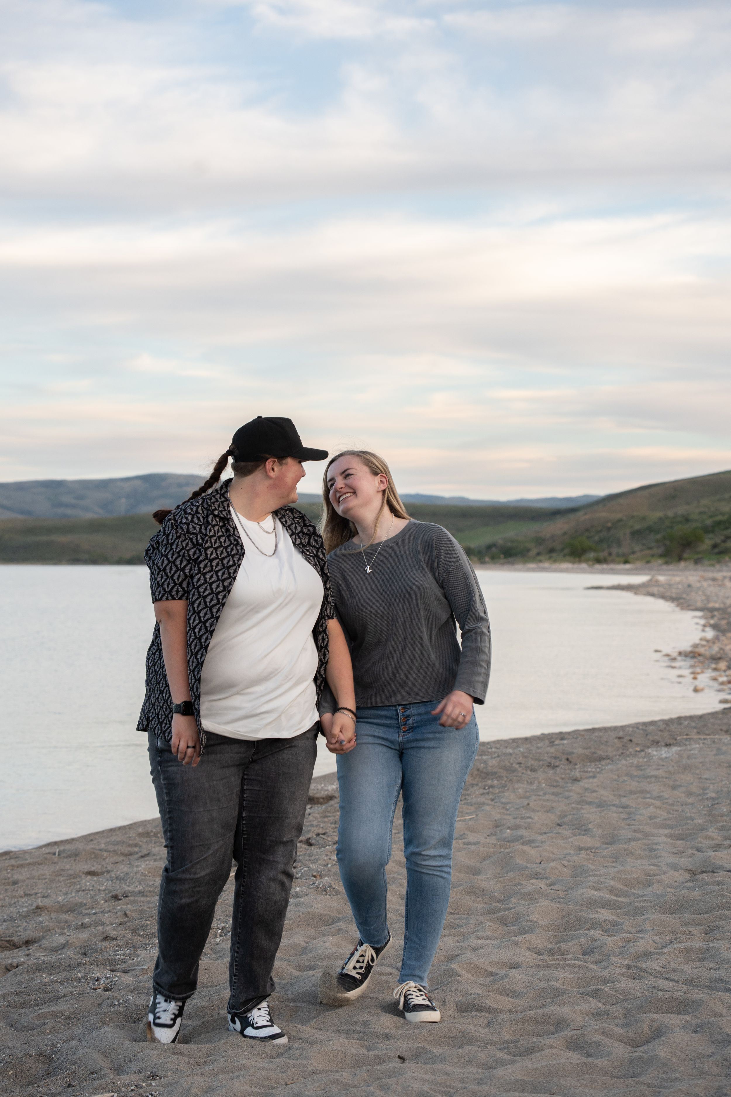 Two women laughing and holding hands while walking along a sandy bear lake beach shoreline at dusk.