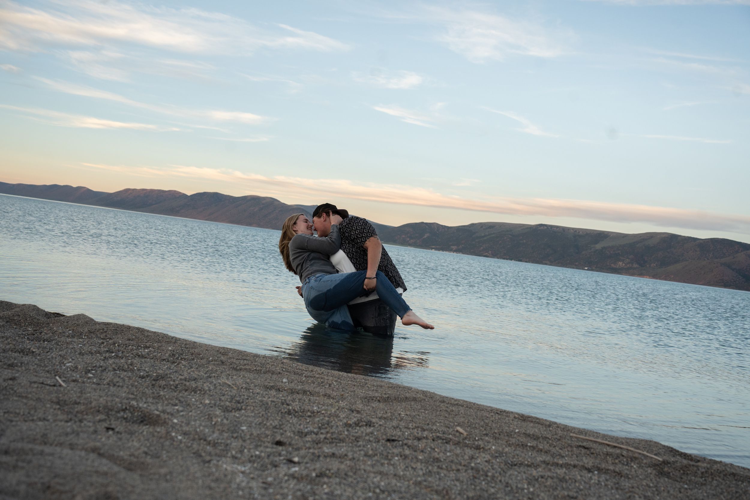 WLW Couple sharing a romantic kiss while standing in shallow bear lake water at a scenic lakeside beach during sunset.