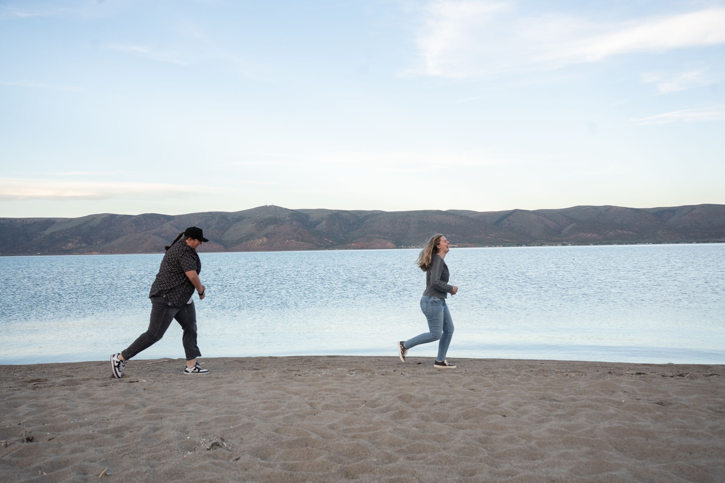 Two people jogging along a sandy beach with calm blue water and mountains in the background.