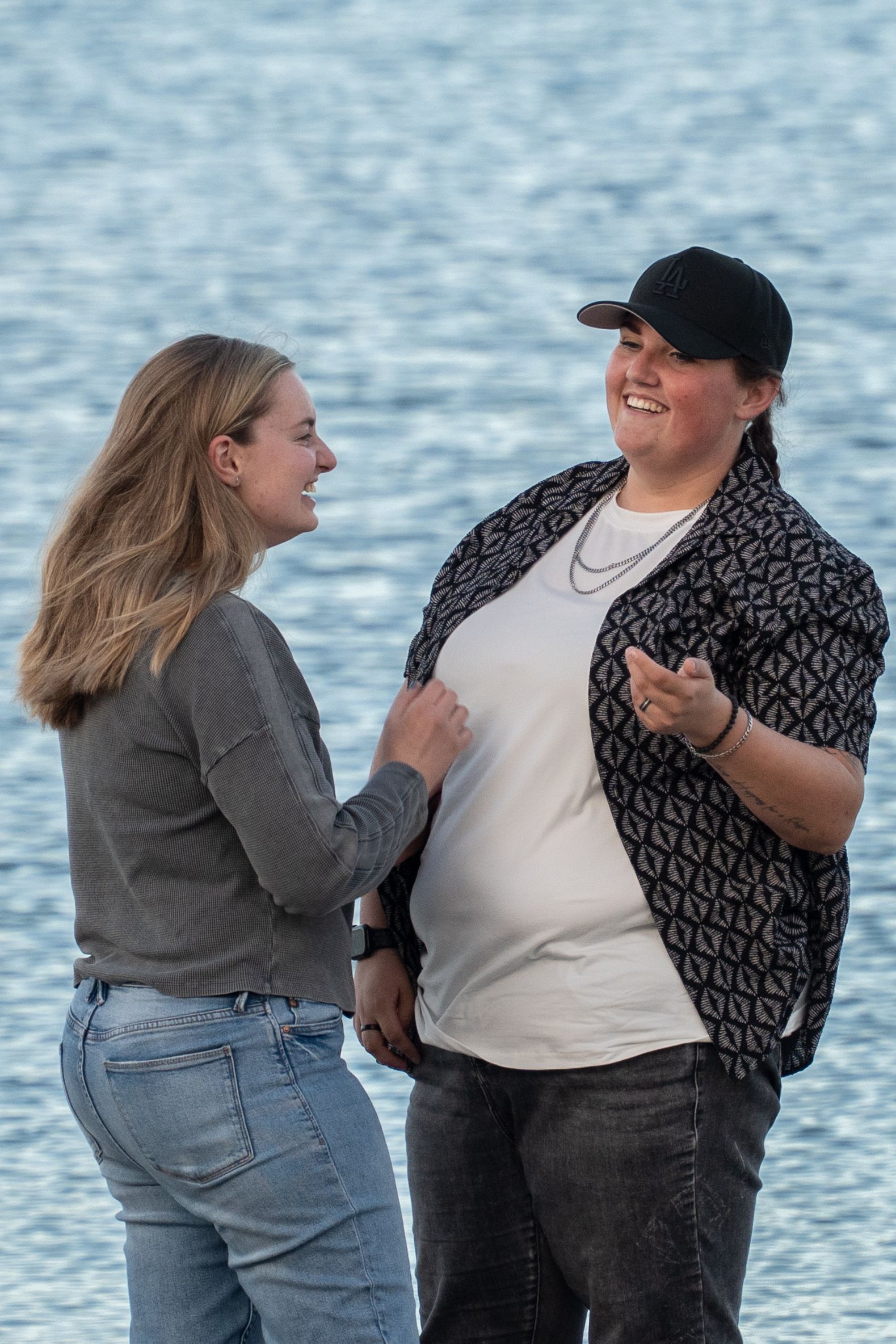 Two people smiling and talking near a shimmering blue waterfront on a sunny day.
