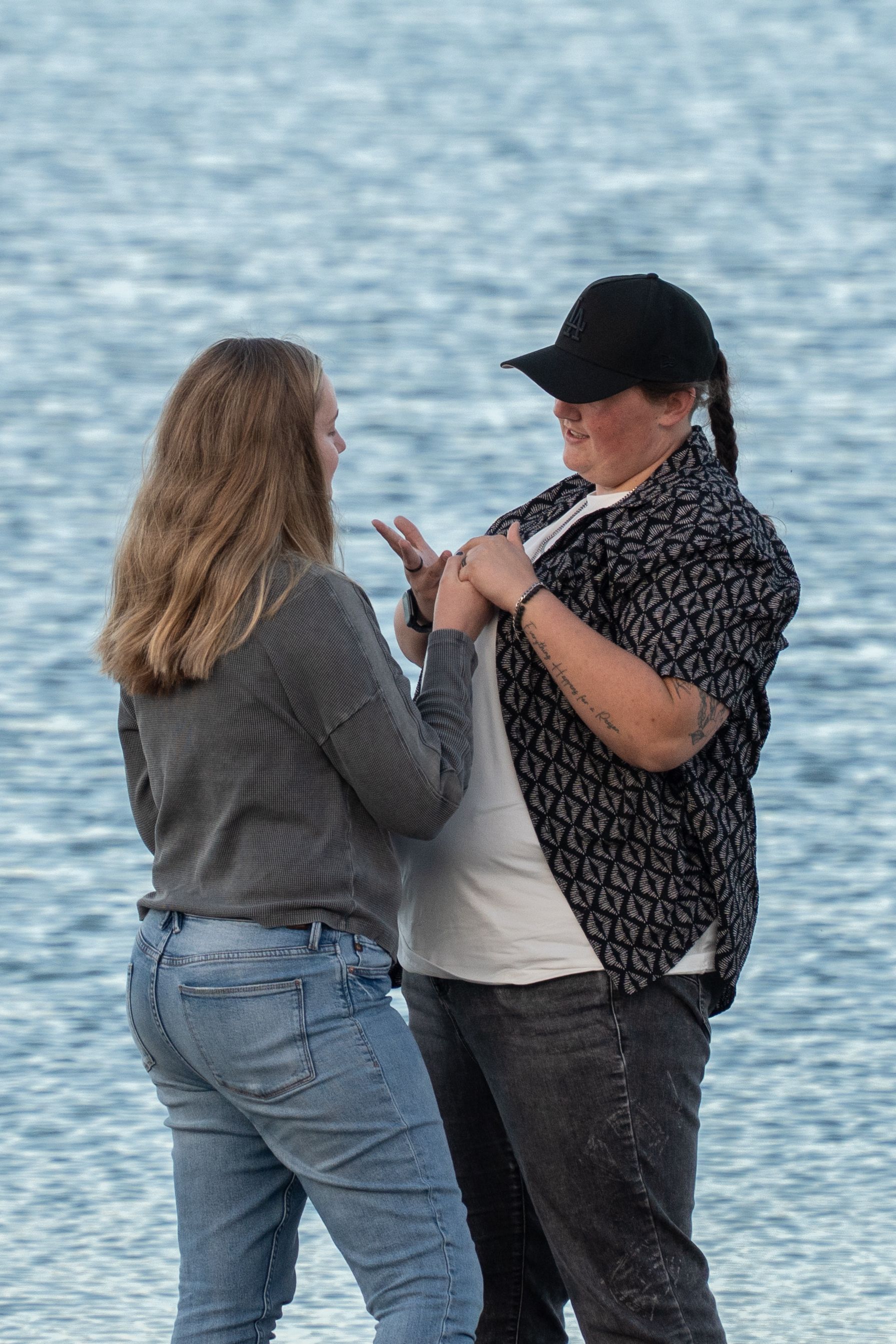 Two women holding hands near a shimmering blue lake, one wearing a black cap and patterned shirt, the other in jeans and gray top.