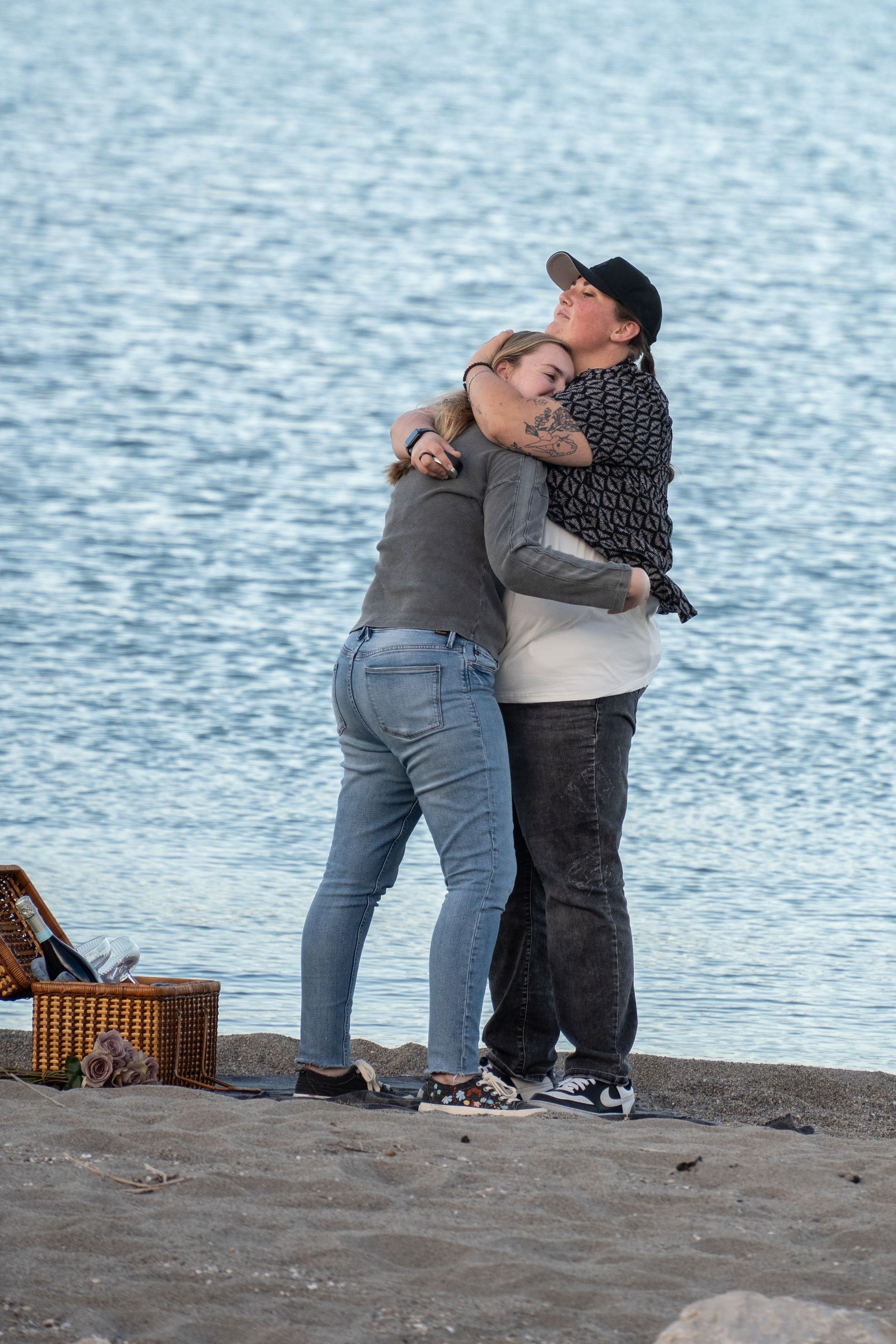 wlw Couple embracing in a warm hug by the waterfront near a picnic basket on rocky shore.