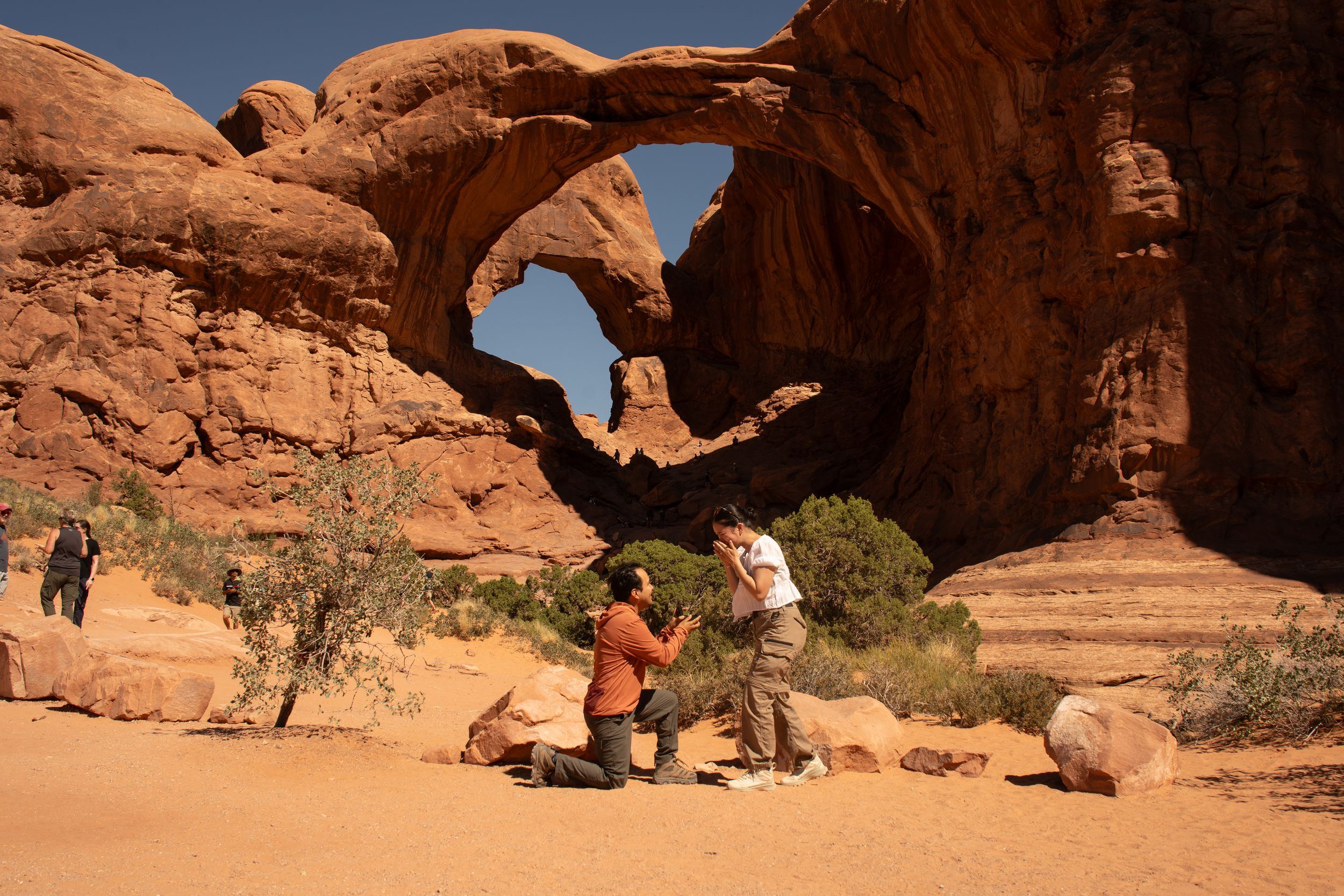 Couple getting engaged beneath Double Arch at Arches National Park, Utah, with red sandstone formations.