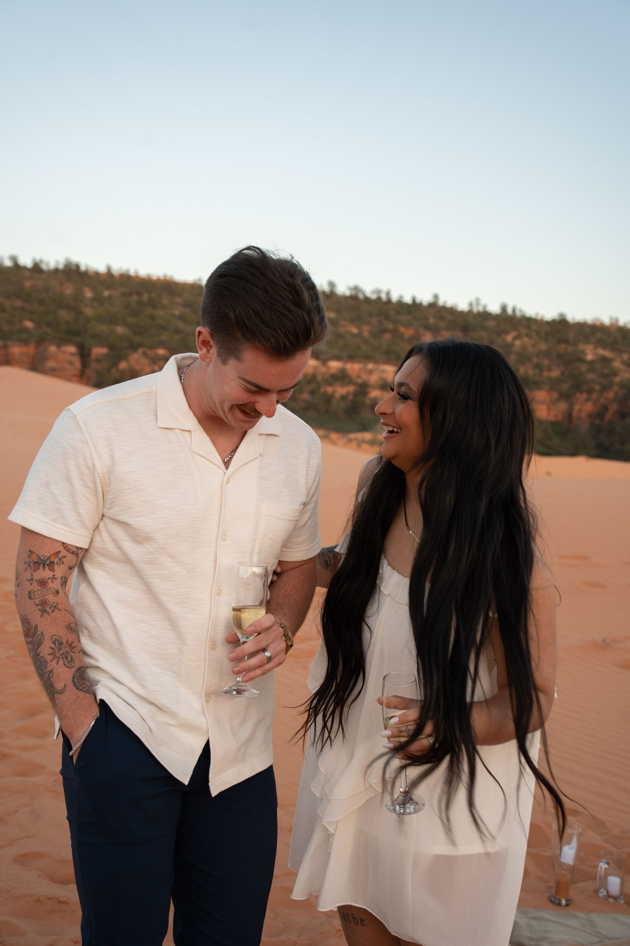 newly engaged Couple holding champagne glasses smiling on red sand dunes at sunset, man with tattoos in white shirt.