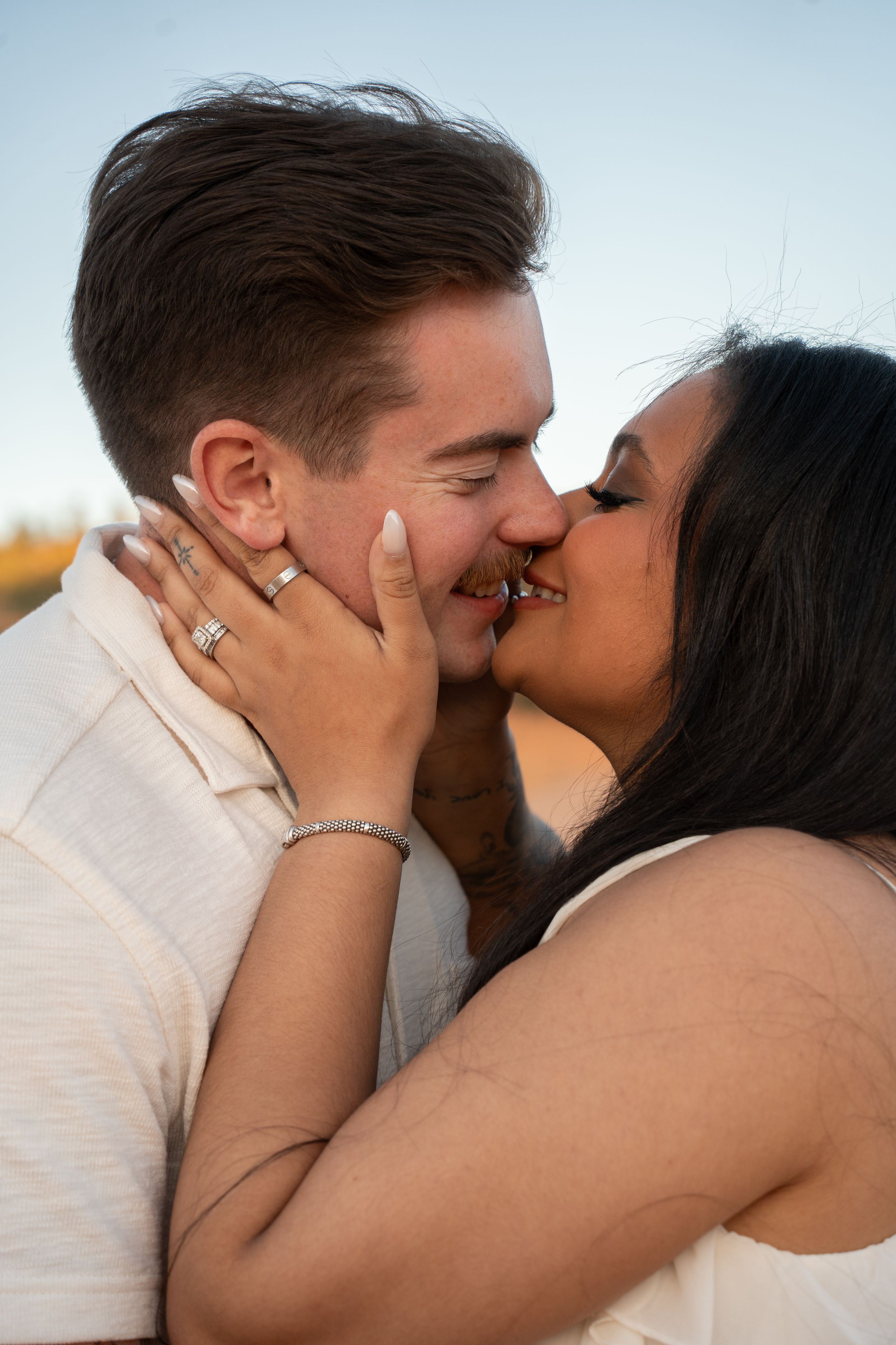 A couple shares a romantic kiss outdoors, with the woman wearing an engagement ring and bracelet.