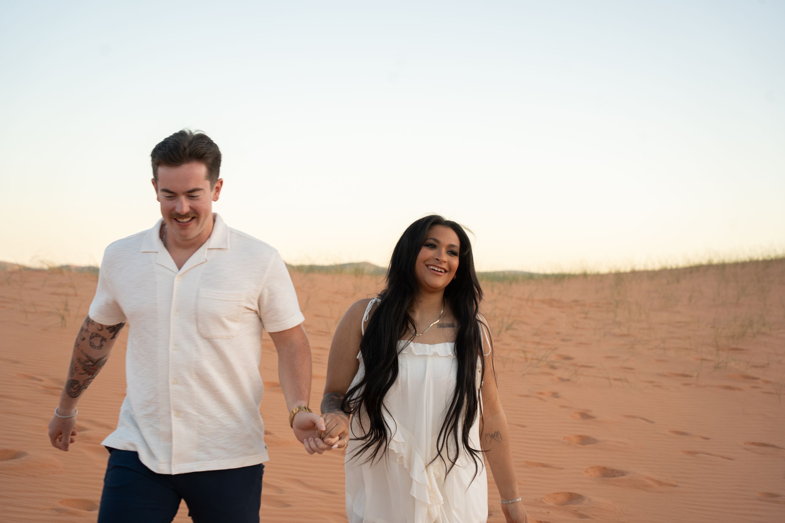 Couple holding hands walking on red sand desert dunes at sunset, man in white shirt and woman in white dress.