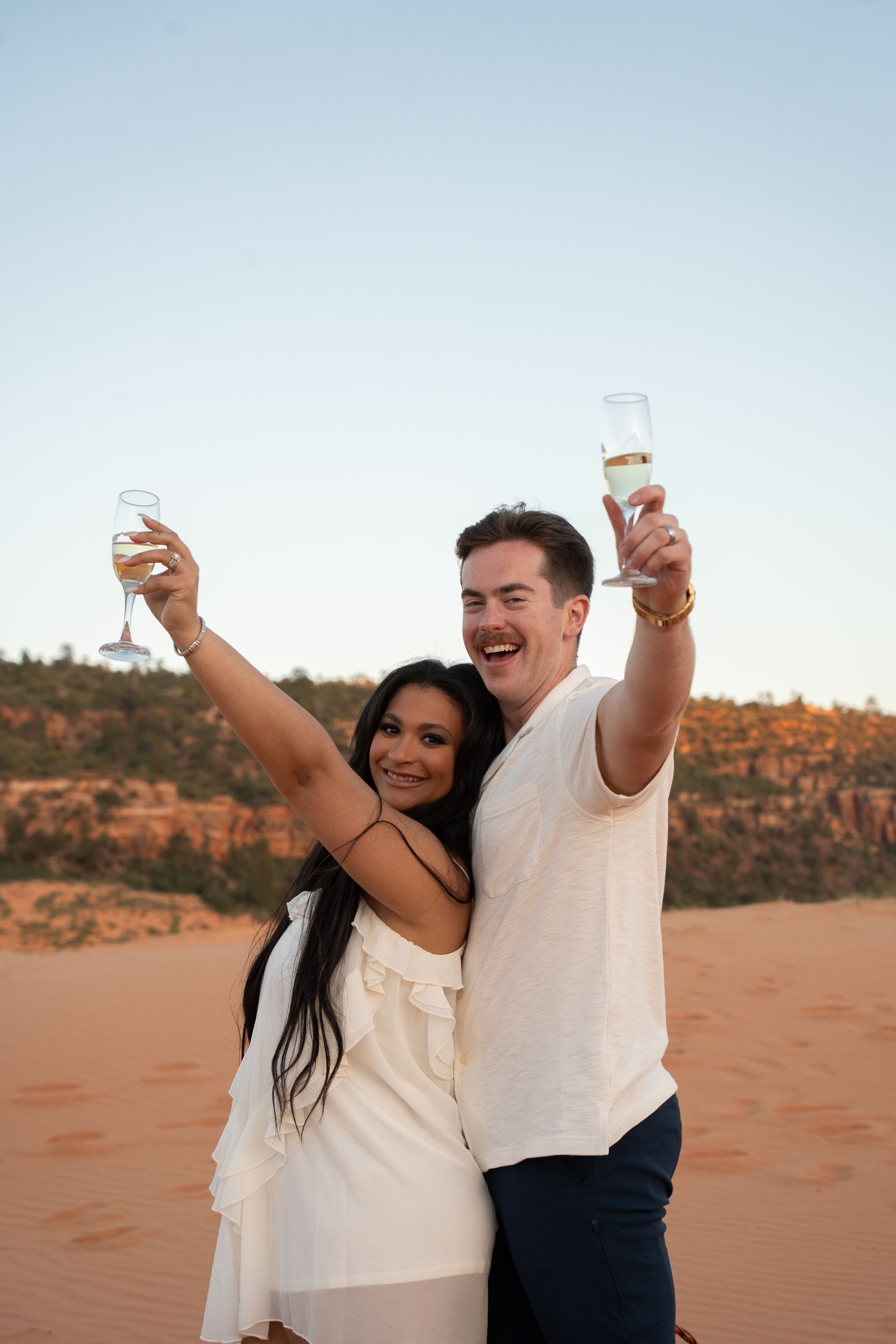 Happy couple celebrating their proposal with champagne glasses raised on kanab pink sand dunes at sunset in white outfits.