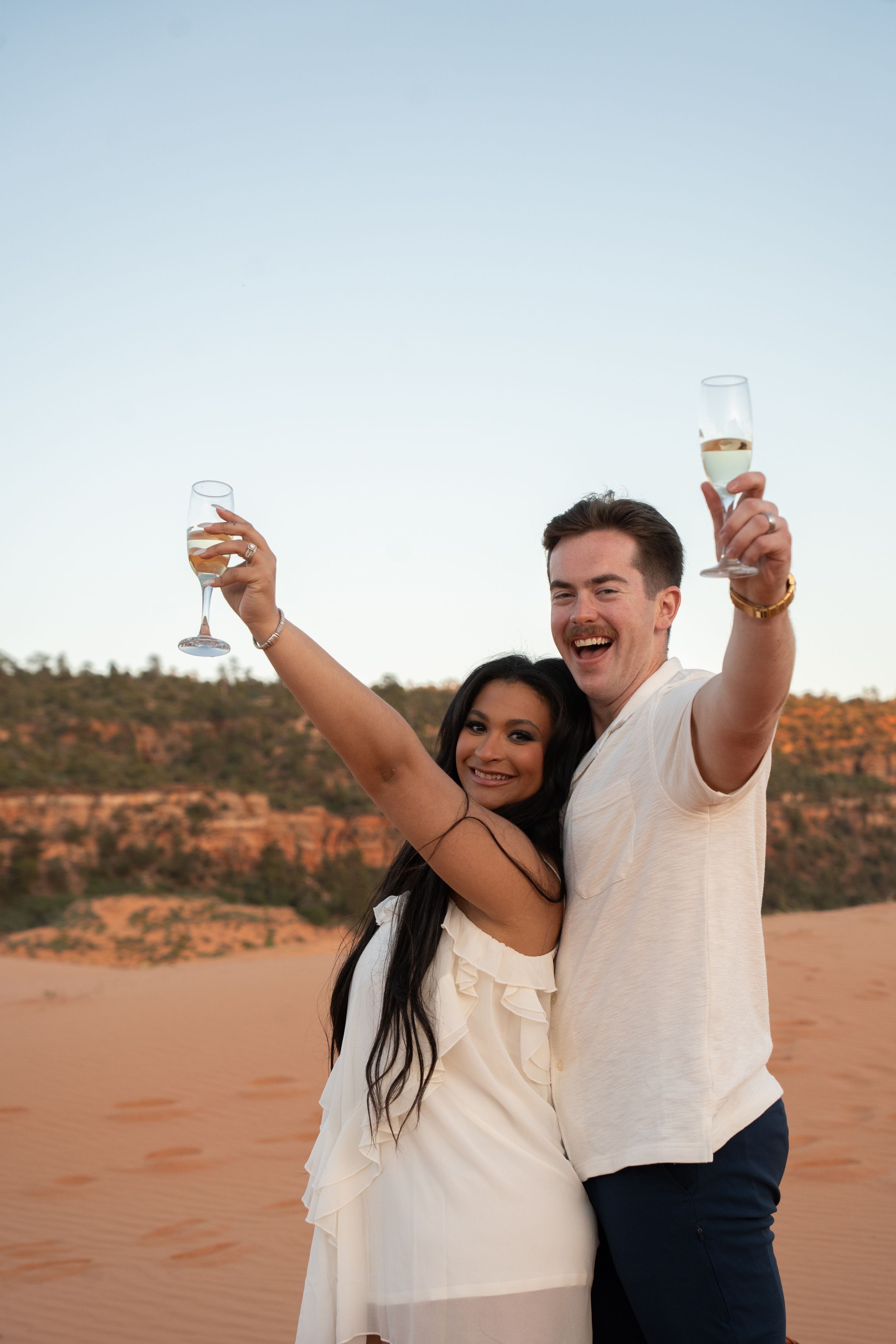 Couple celebrating outdoors in desert landscape, raising champagne glasses and smiling, wearing white outfits at sunset.