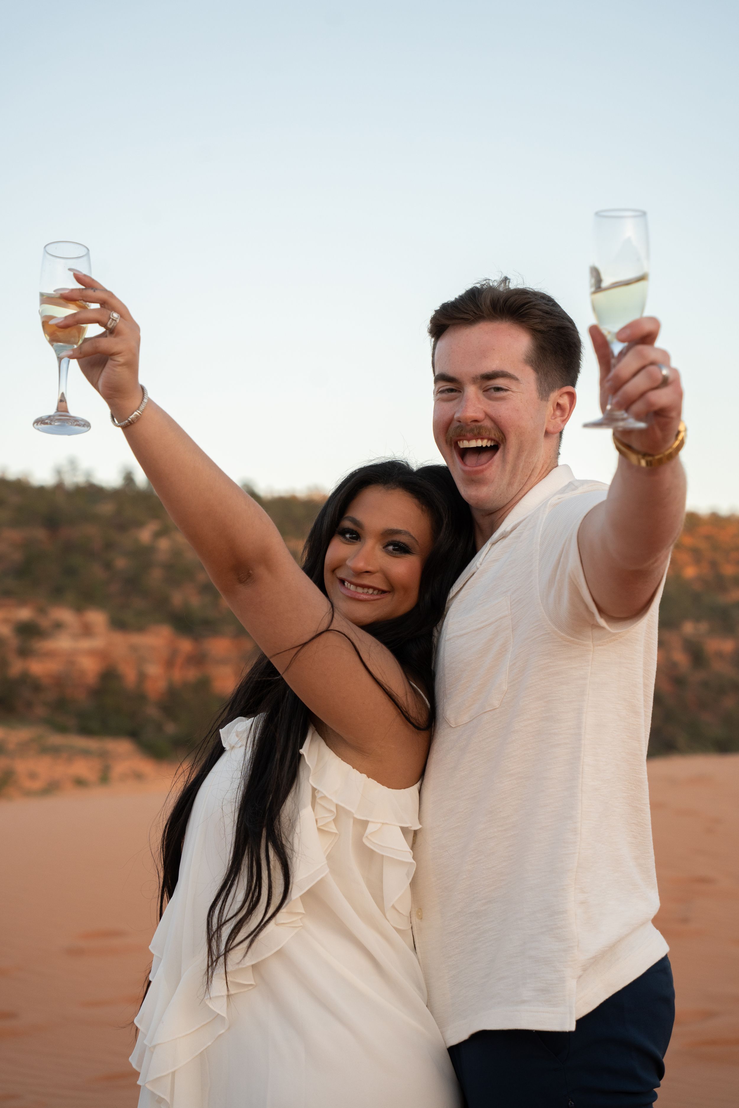 Couple in white outfits toasting with champagne glasses in coral pink sand dunes of kanab