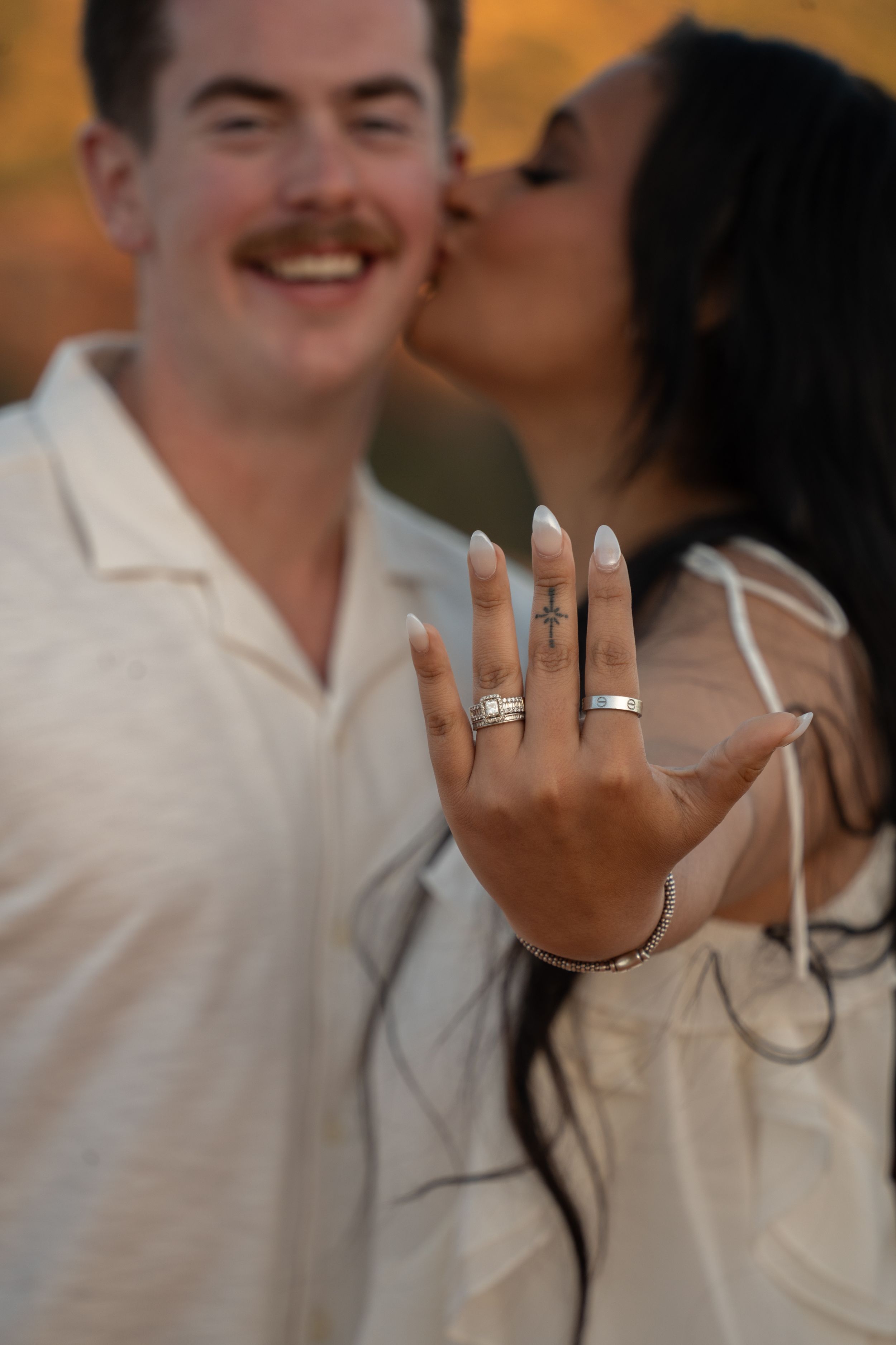 Couple in white outfits showing off engagement rings, woman kissing smiling man outdoors at golden hour.