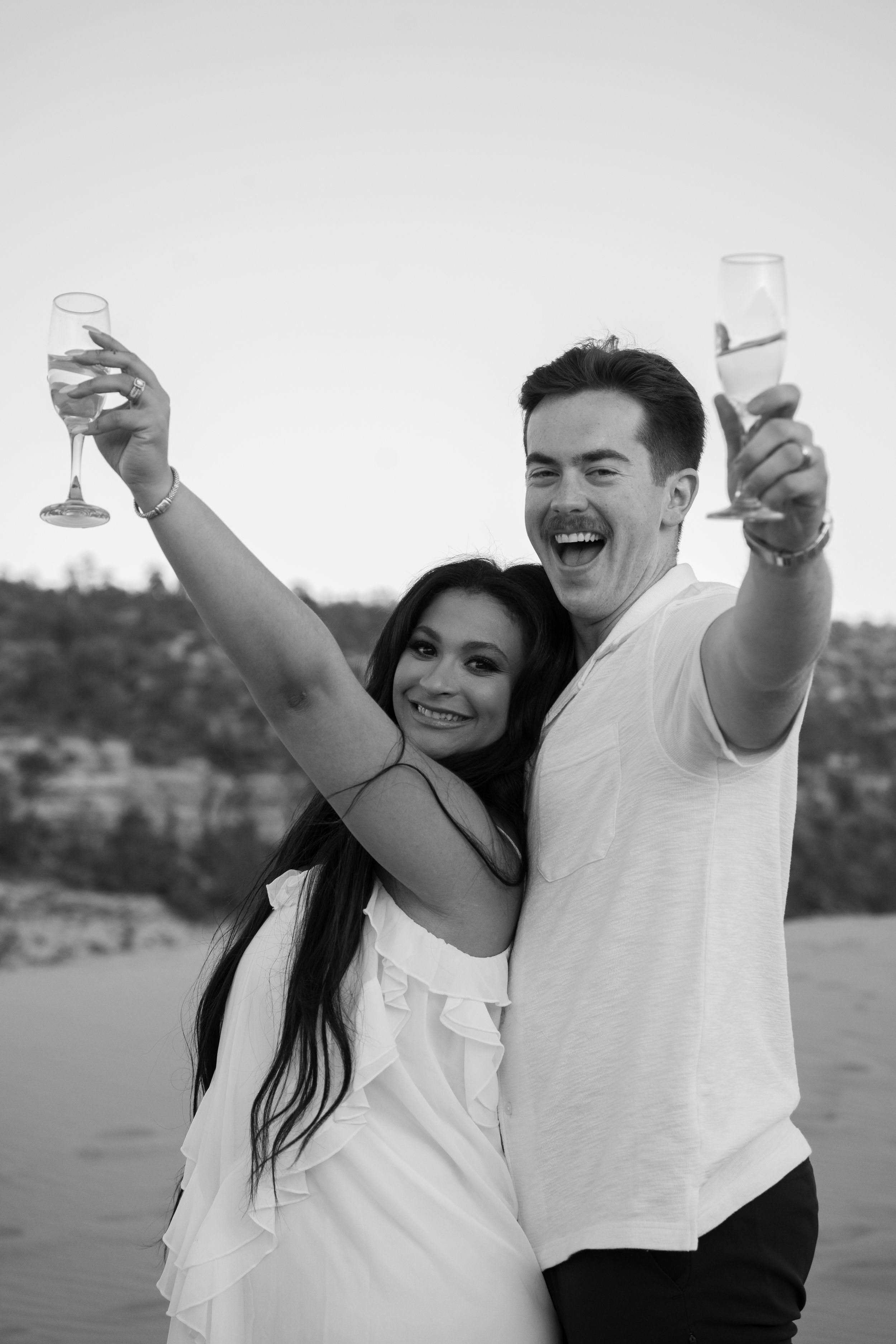Black and white photo of a joyful couple raising champagne glasses in a celebratory toast at kanab pink sand dunes.