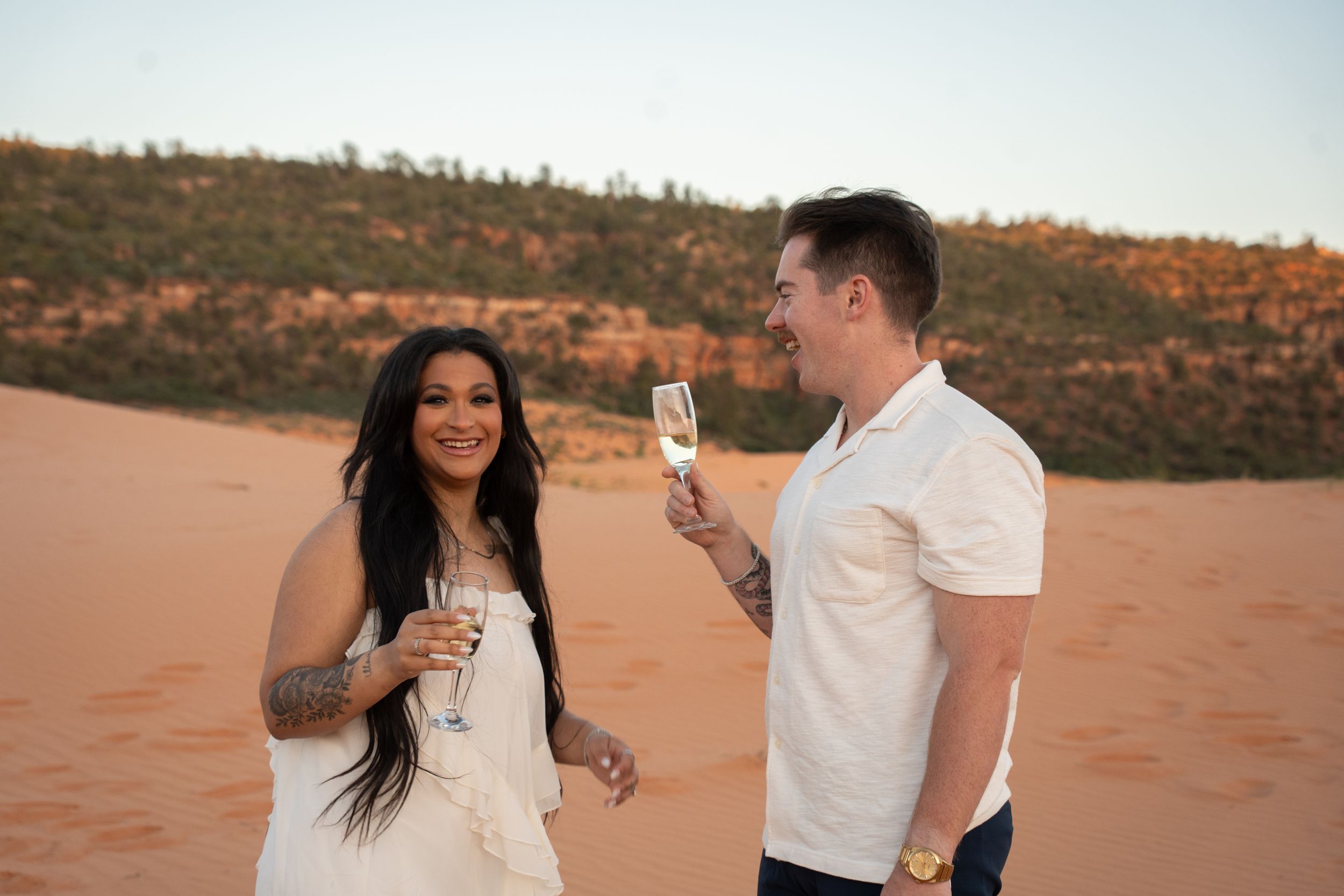 Couple toasting with champagne glasses on coral pink sand dunes at sunset, woman with tattoos in white dress, man in white shirt.