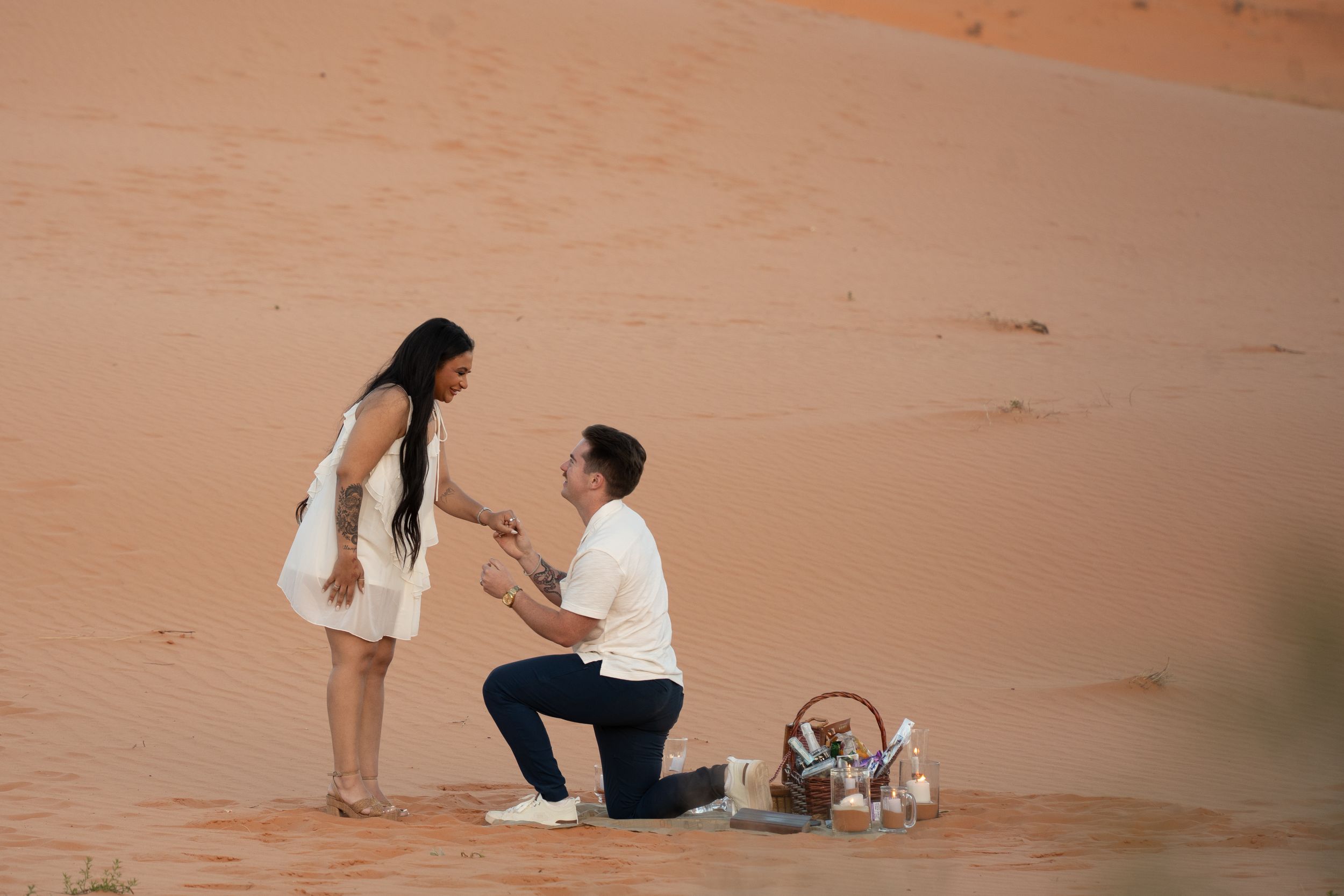 Man proposing on one knee to woman in white dress on sandy desert dunes with romantic picnic basket nearby.