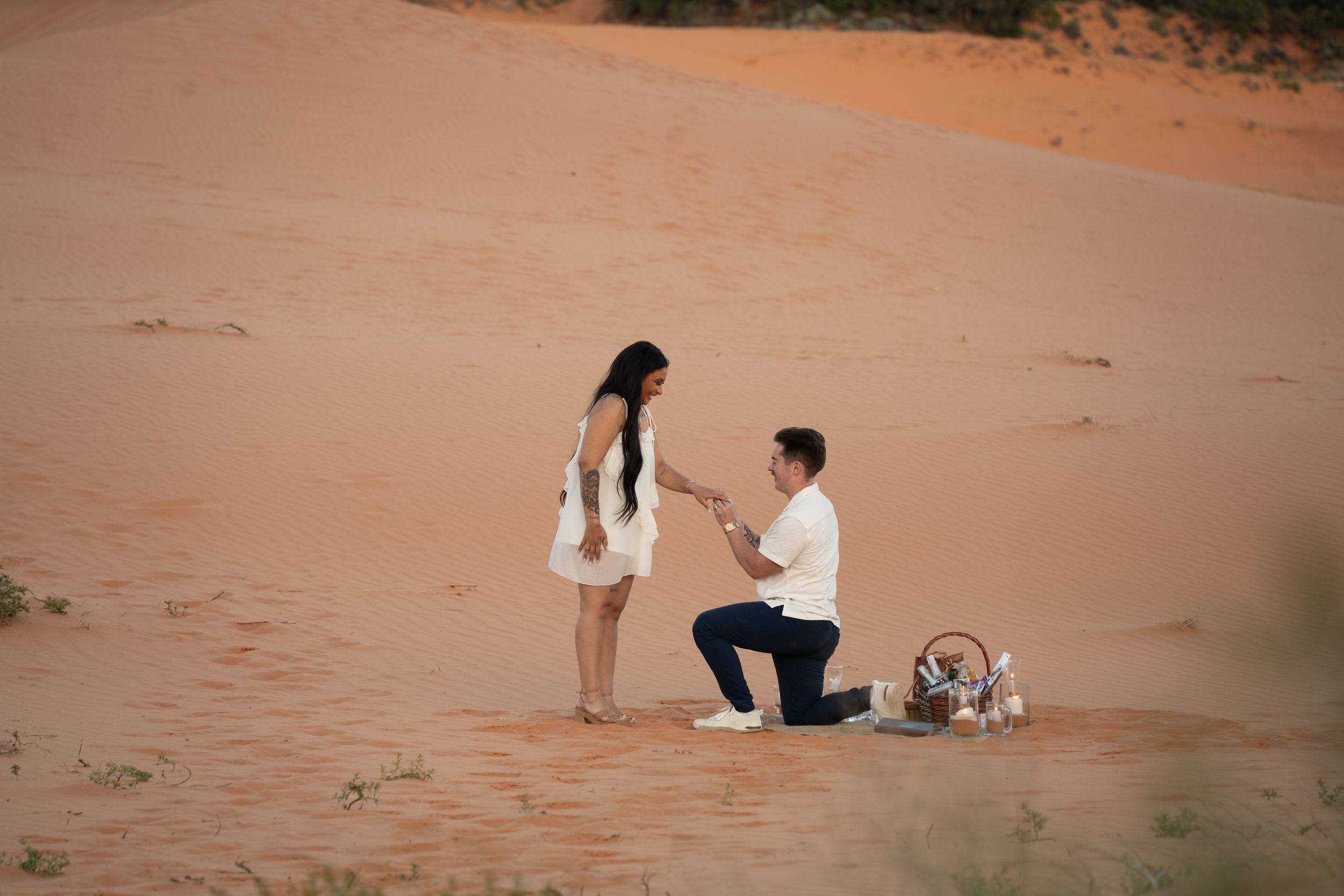 Man proposing to woman on coral pink sand desert dunes, kneeling with ring while picnic basket sits nearby.