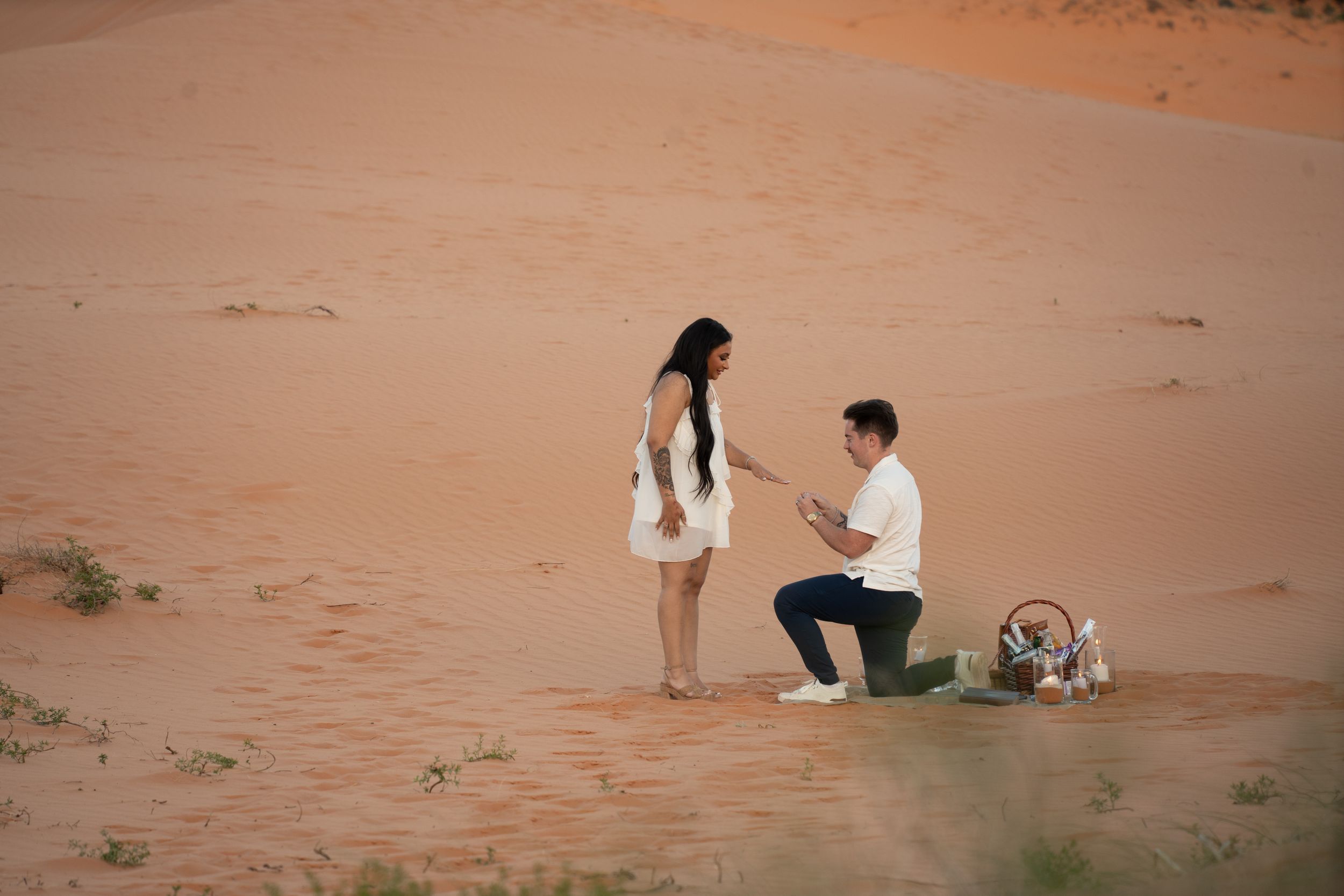 Man proposing to woman on one knee in a stunning red sand desert landscape with a picnic basket nearby.