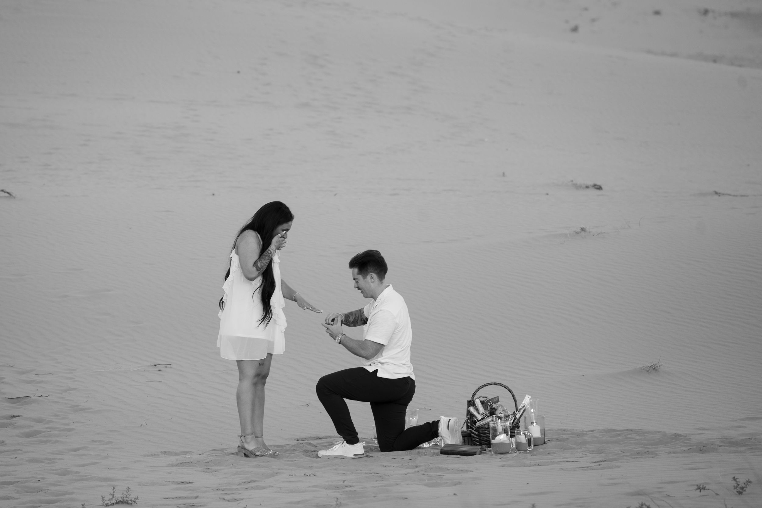 Man proposing on one knee to woman on sandy pink dunes in romantic black and white photography.