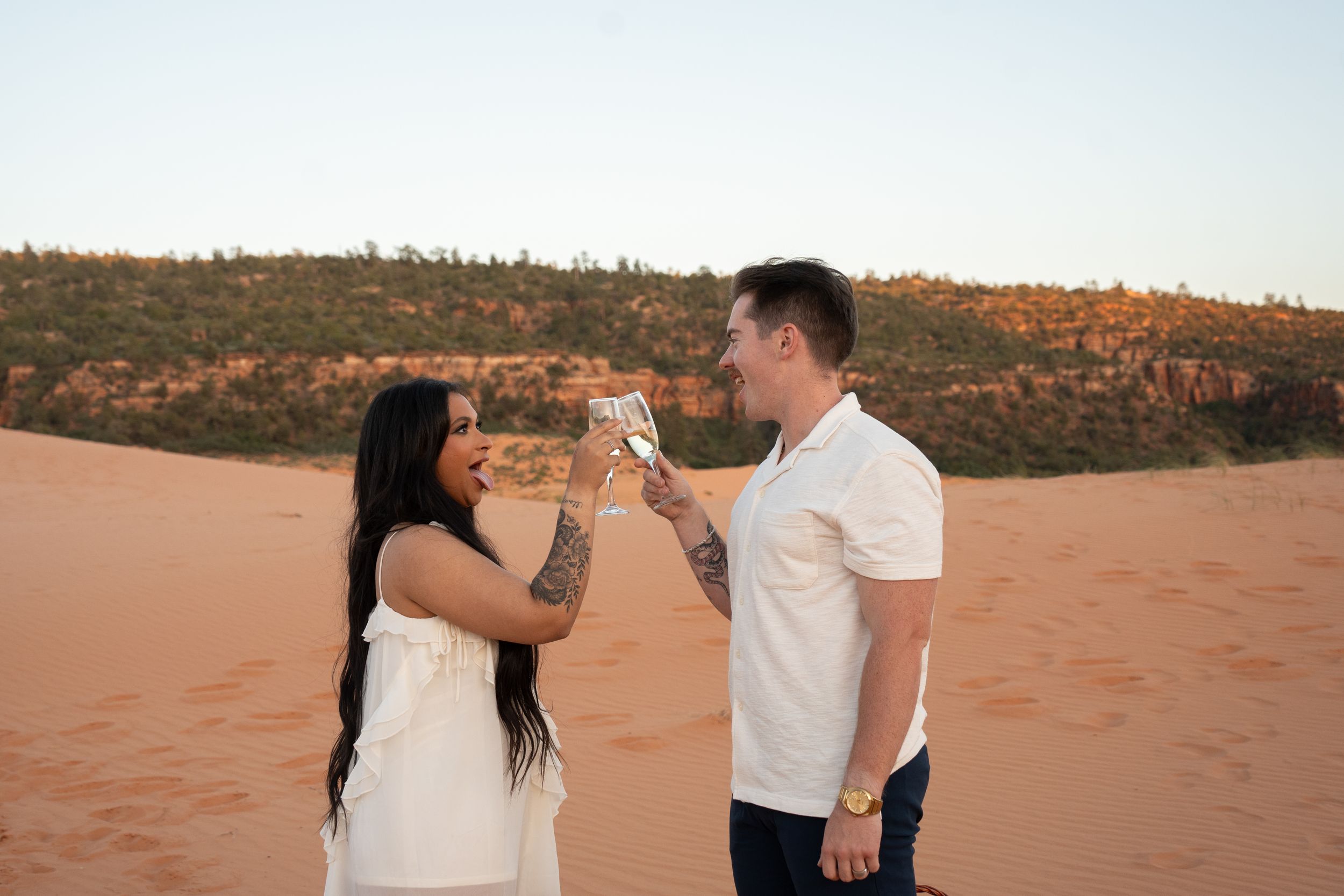 Couple toasting with champagne flutes to celebrate a proposal on red sand dunes with green trees and rocky cliffs in background.