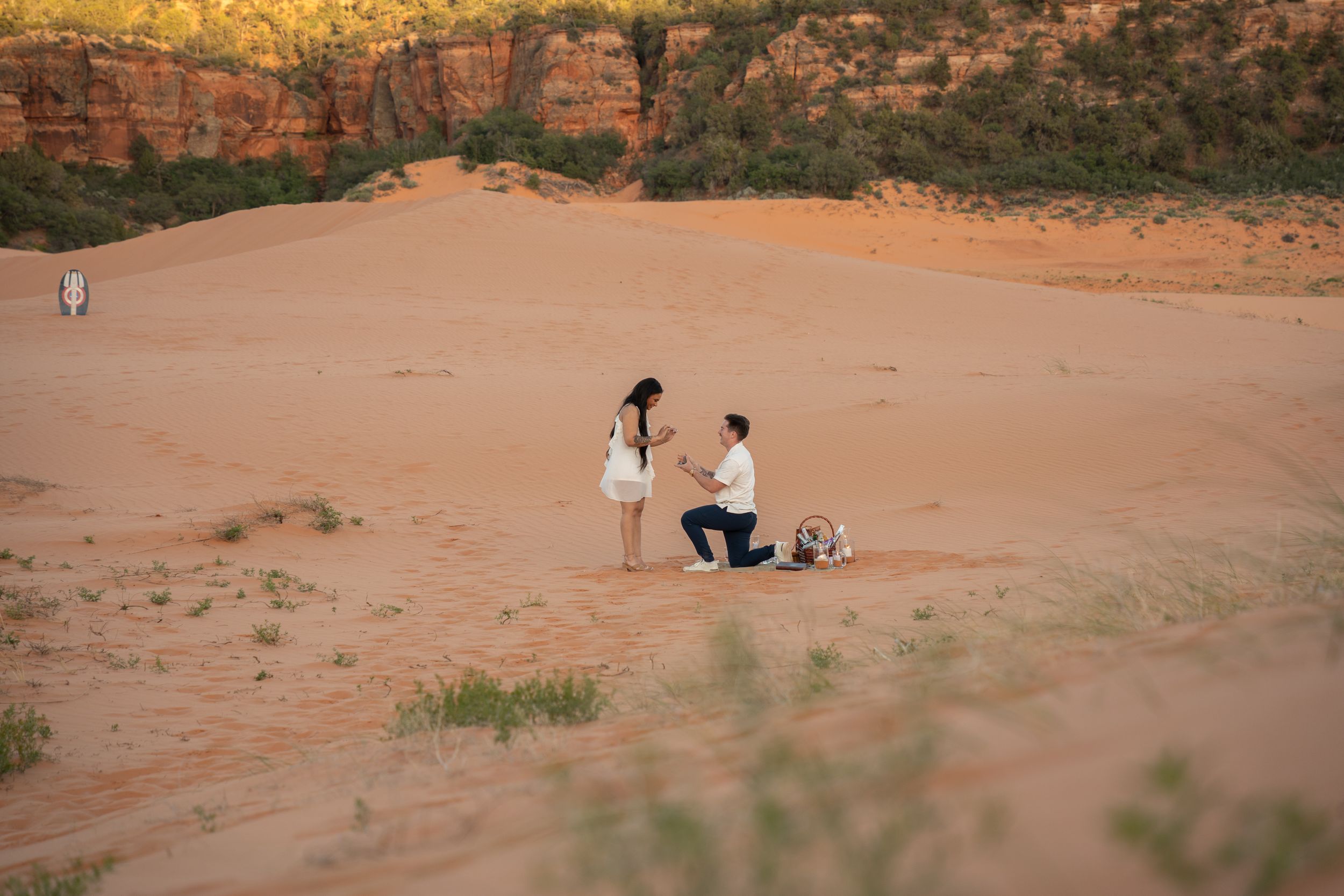 Man proposes on one knee to woman on red sand dunes in desert landscape with red rock cliffs backdrop.