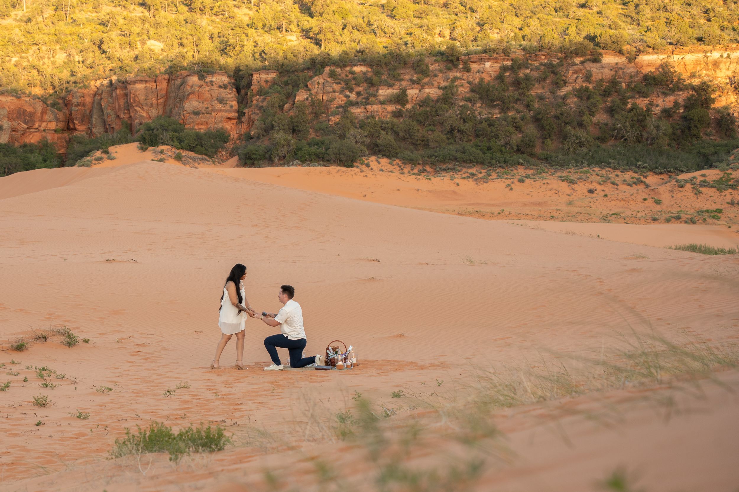 Man proposing to woman on red sand dunes with lush green hillside backdrop at golden hour.