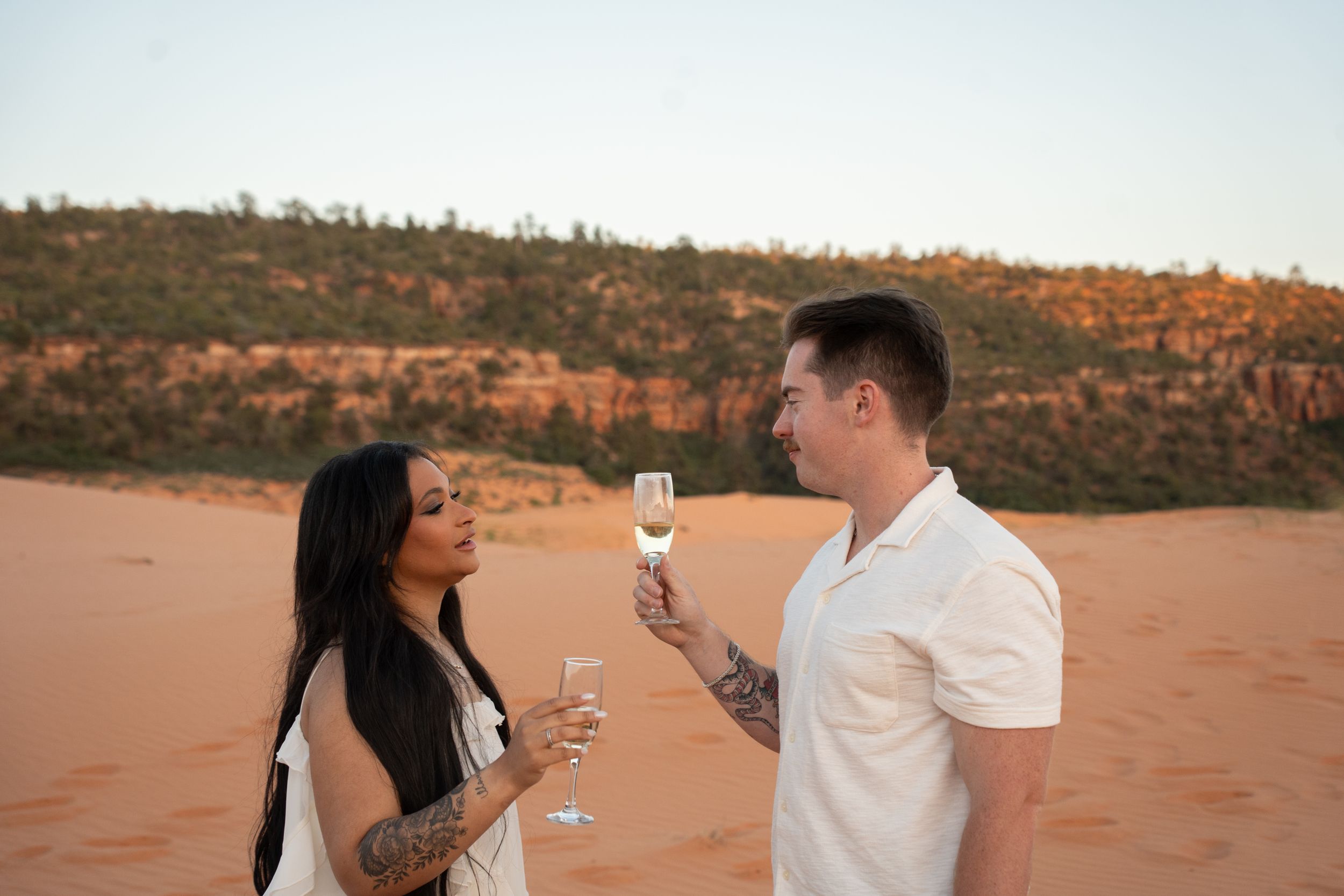 Couple toasting with champagne glasses on red sand desert landscape at sunset, both with tattoos.