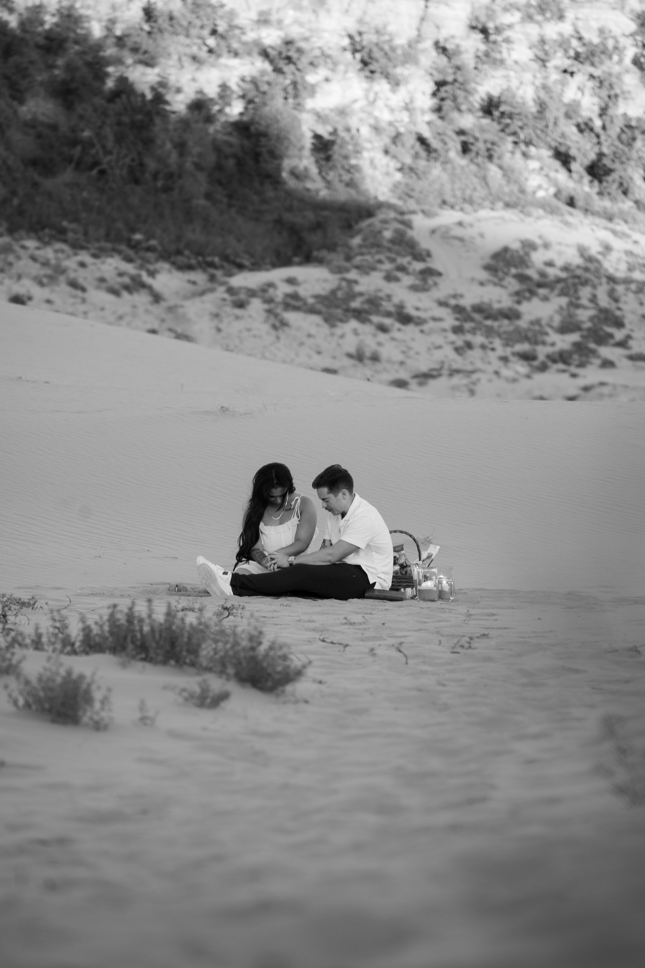 Black and white photo of a couple praying together on sandy dunes, sharing a peaceful moment in nature before a proposal
