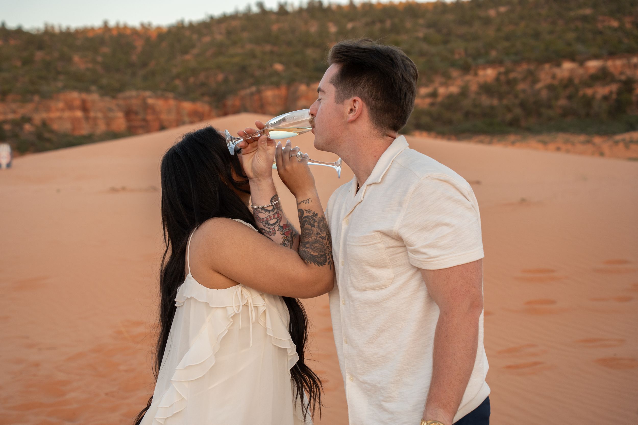 Couple sharing a champagne toast on red sand dunes at sunset in a romantic outdoor celebration of their engagement