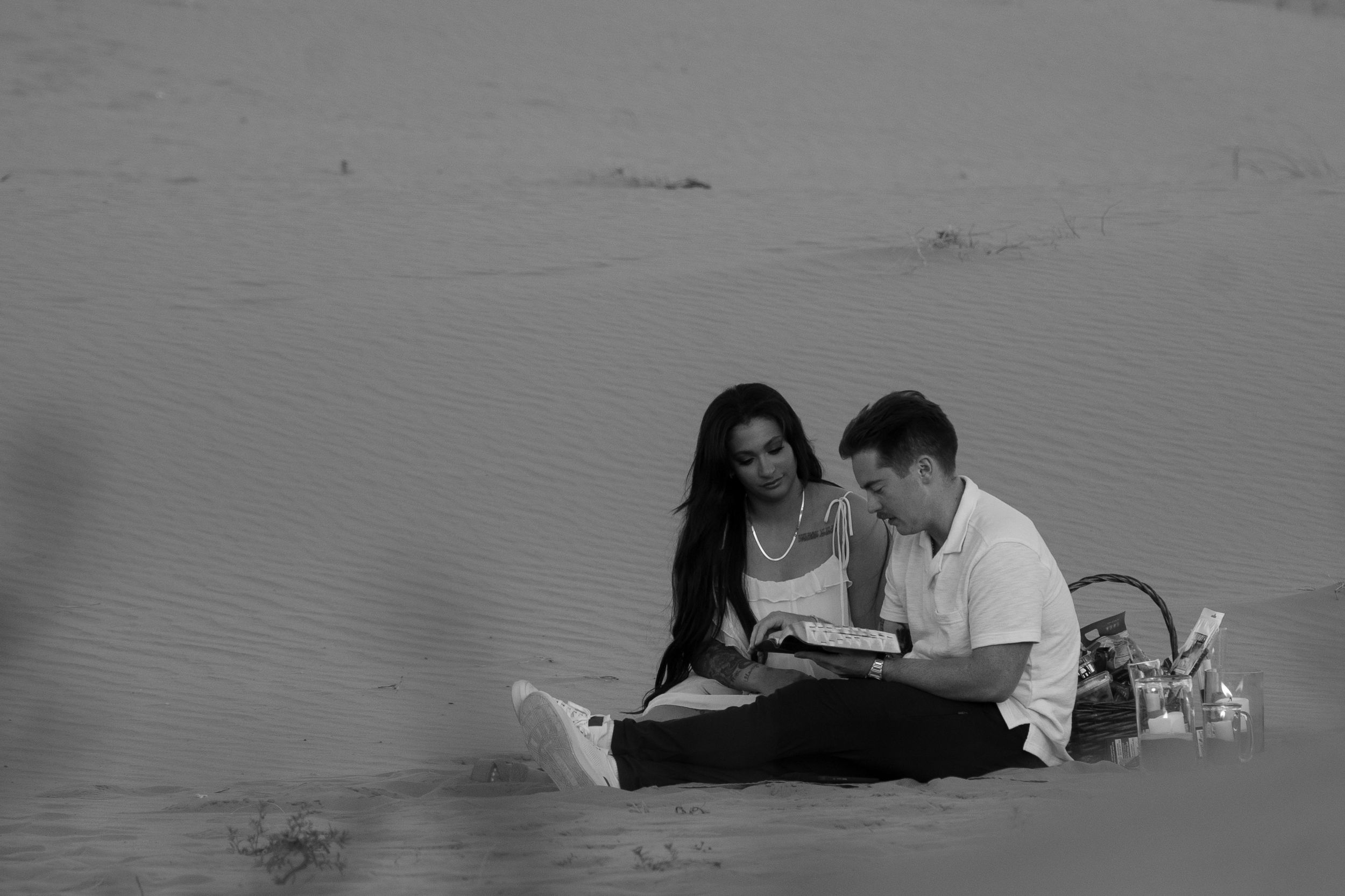Black and white photo of a couple sitting together on sand dunes, sharing an intimate moment reading the bible with a picnic basket nearby.