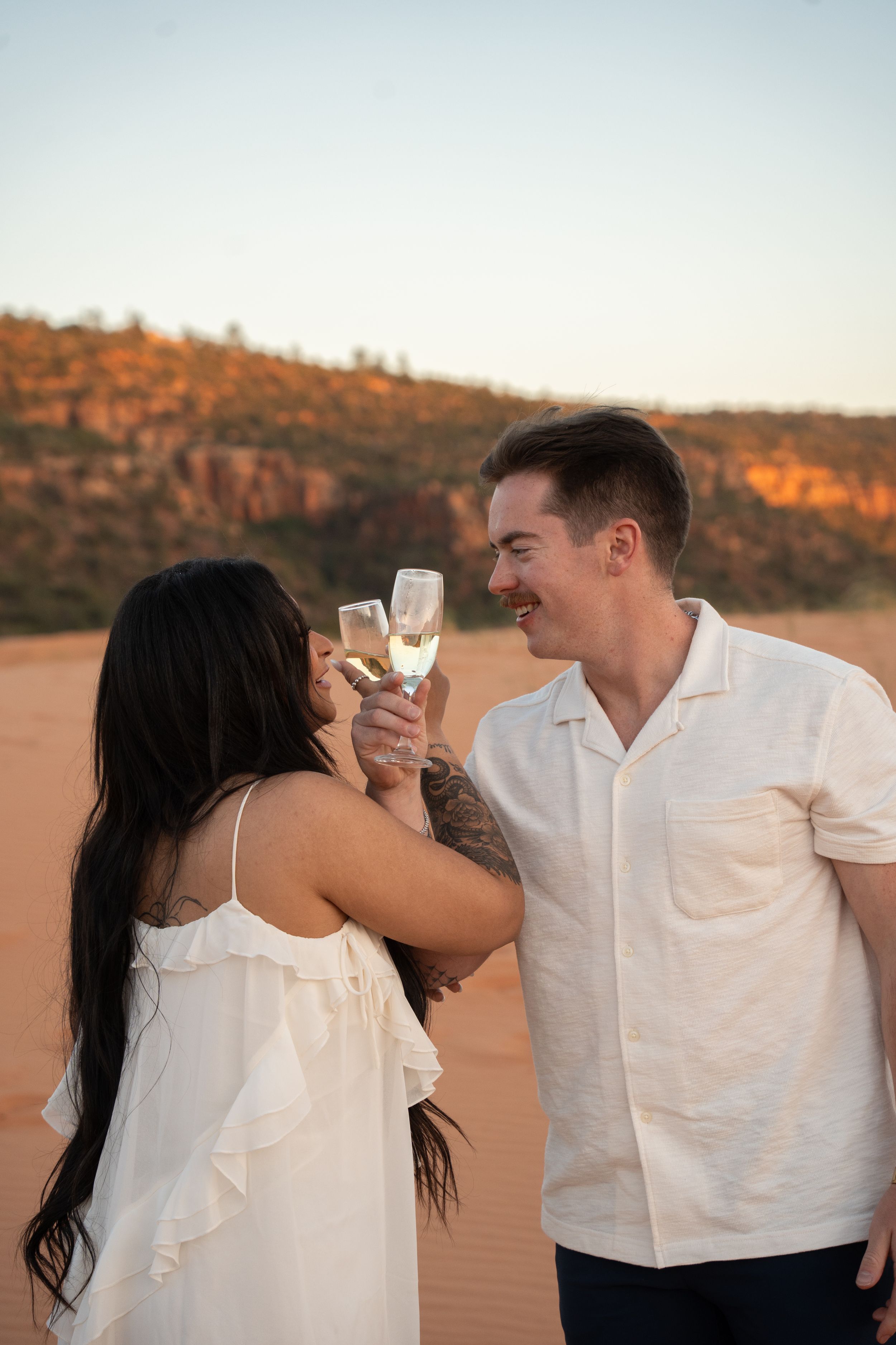 Couple in white outfits toasting with champagne glasses on pink sand dunes at sunset.